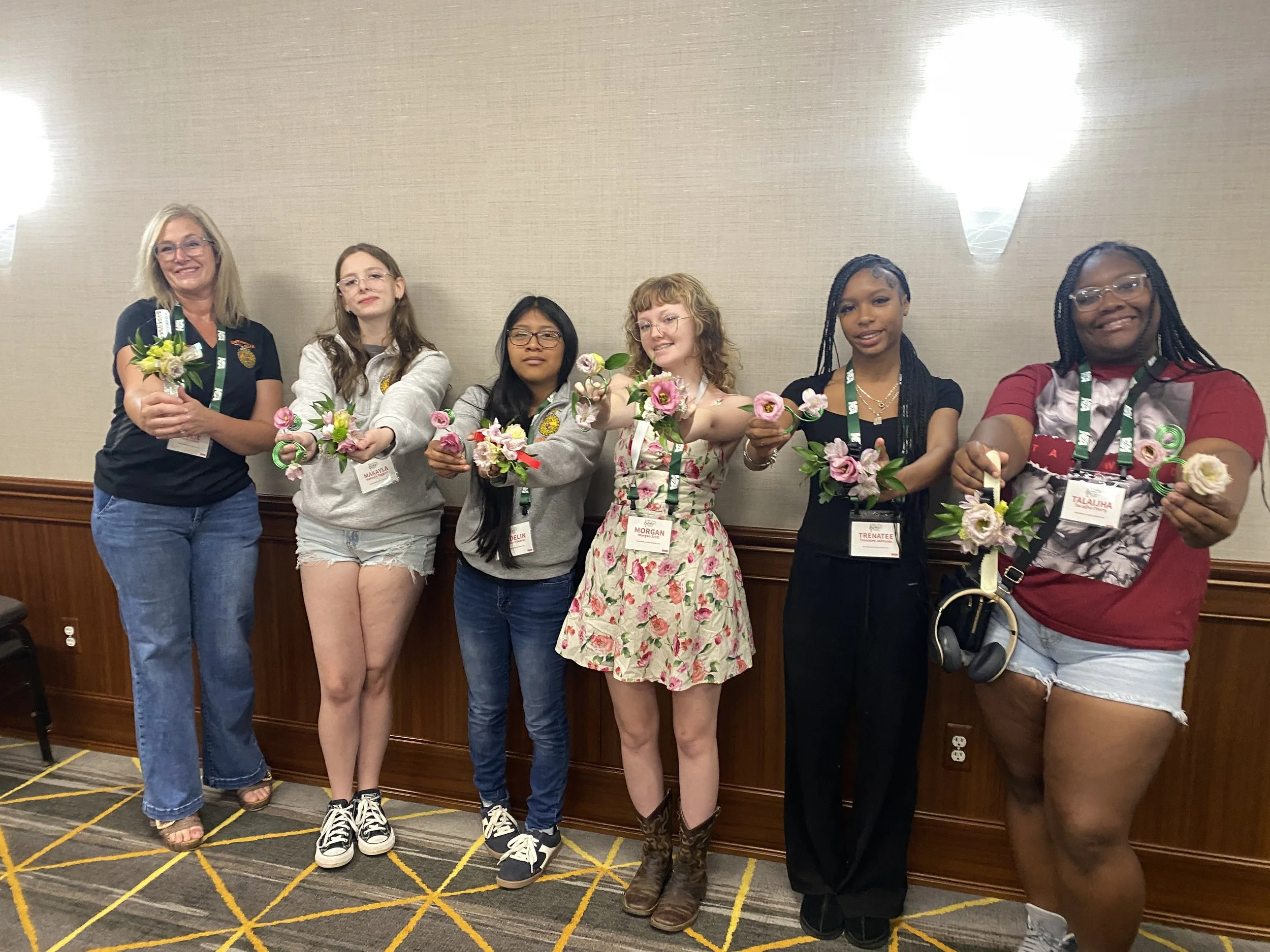 A group of six women standing indoors against a beige wall, holding bouquets of pink and yellow flowers, wearing conference badges, smiling, with a patterned carpet on the floor.