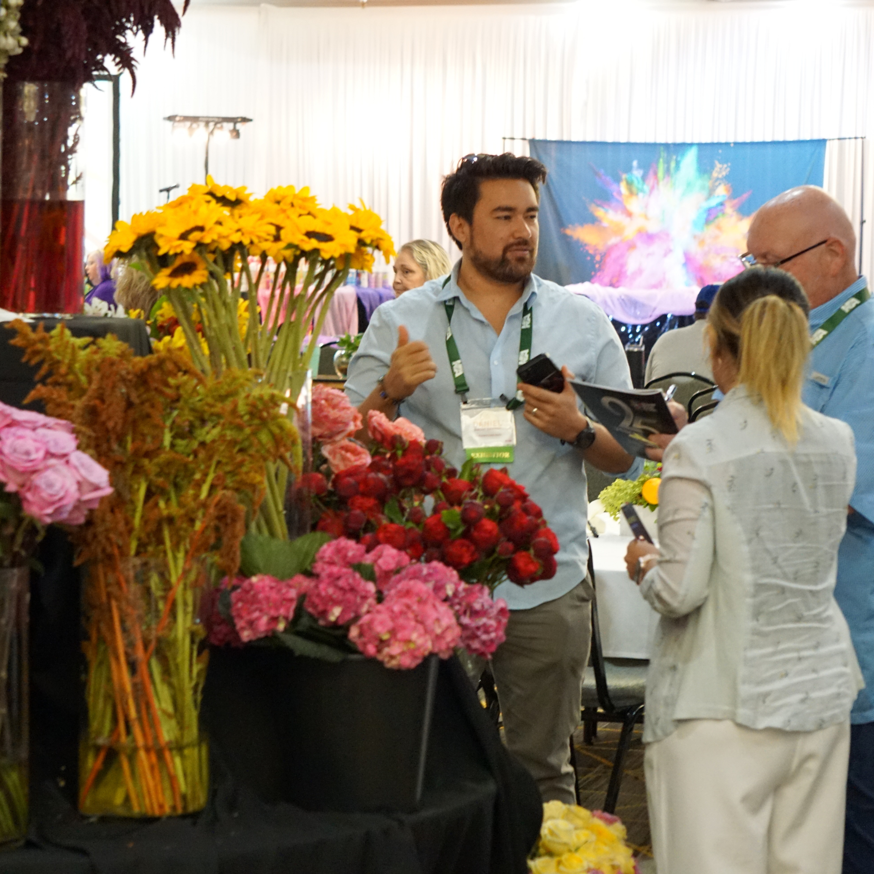 People gathered at an indoor event, engaging in conversation, with a display of colorful flowers and a vibrant, splashy backdrop in the background.