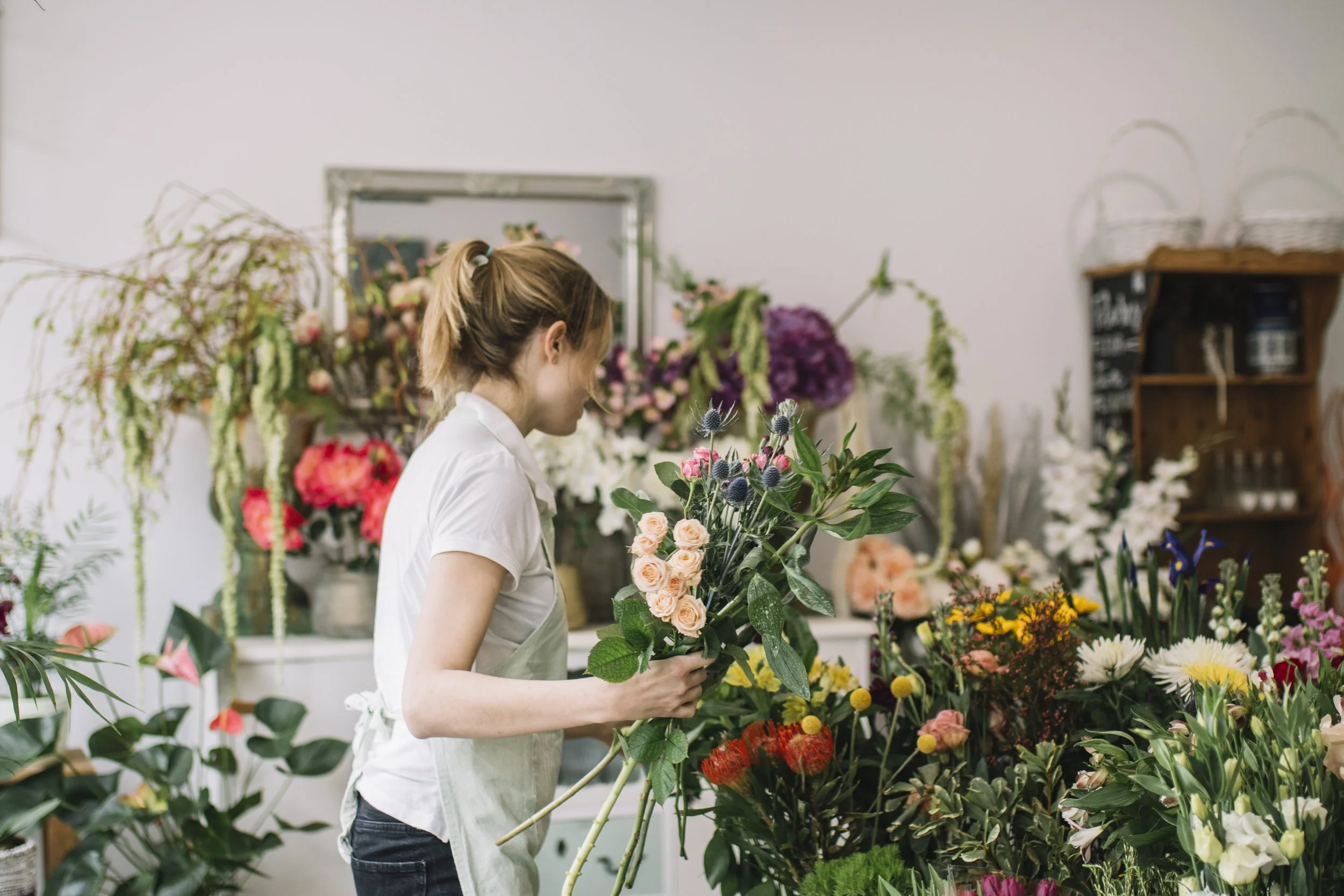 A woman with light brown hair tied back in a ponytail, wearing a white shirt and apron, arranging flowers in a flower shop filled with various colorful flowers and plants.