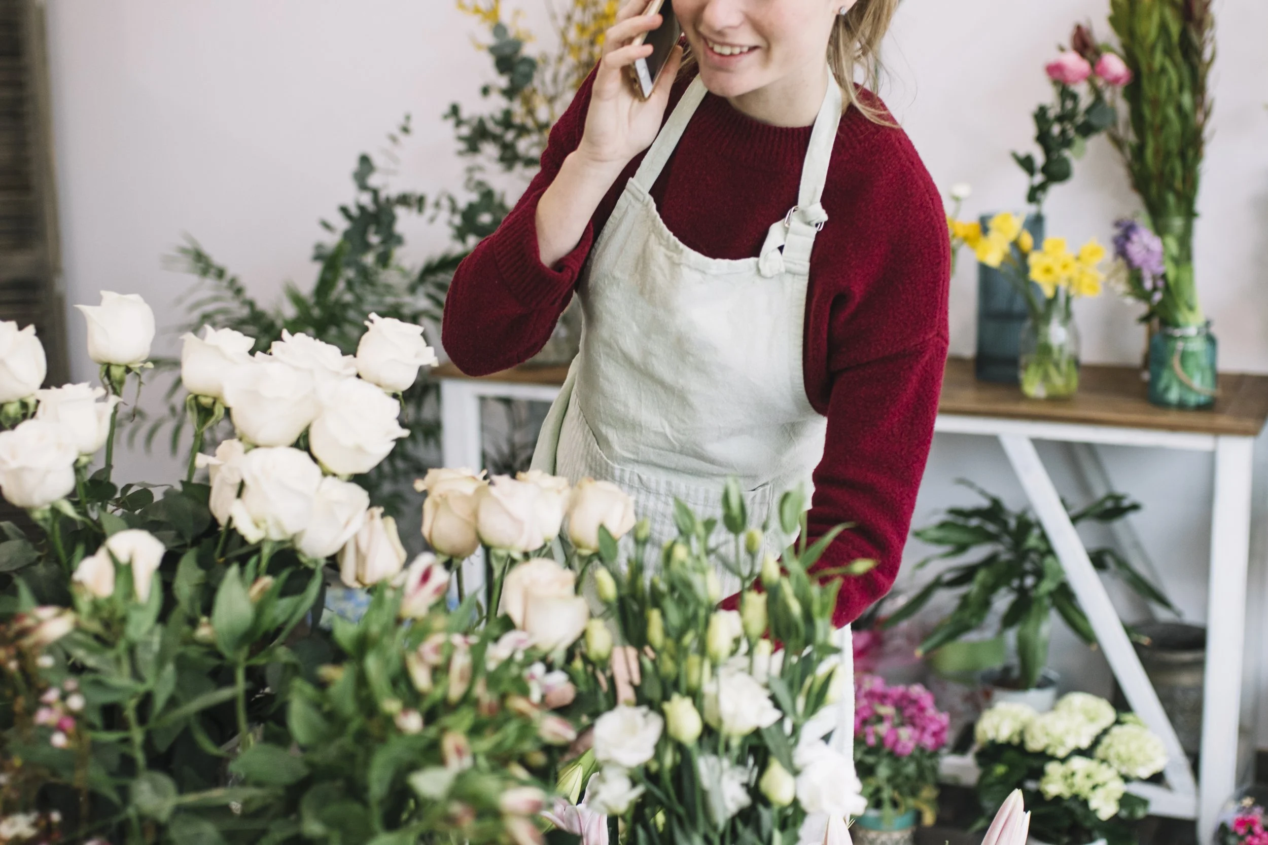 A woman smiling, talking on a phone, and handling flowers in a flower shop. She is wearing a red sweater and a white apron, surrounded by colorful flowers in vases and baskets.