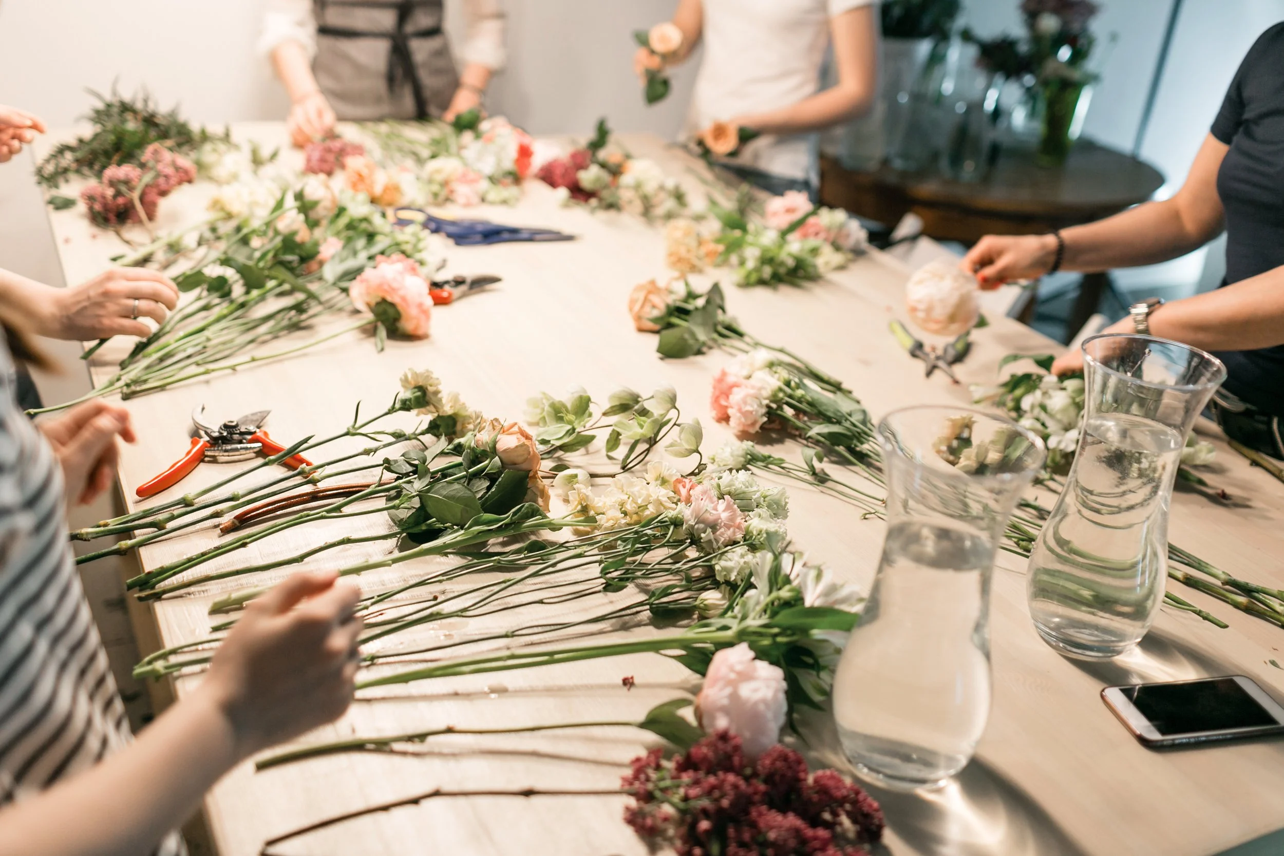 People arranging pink and white flowers on a long table with scissors and vases.