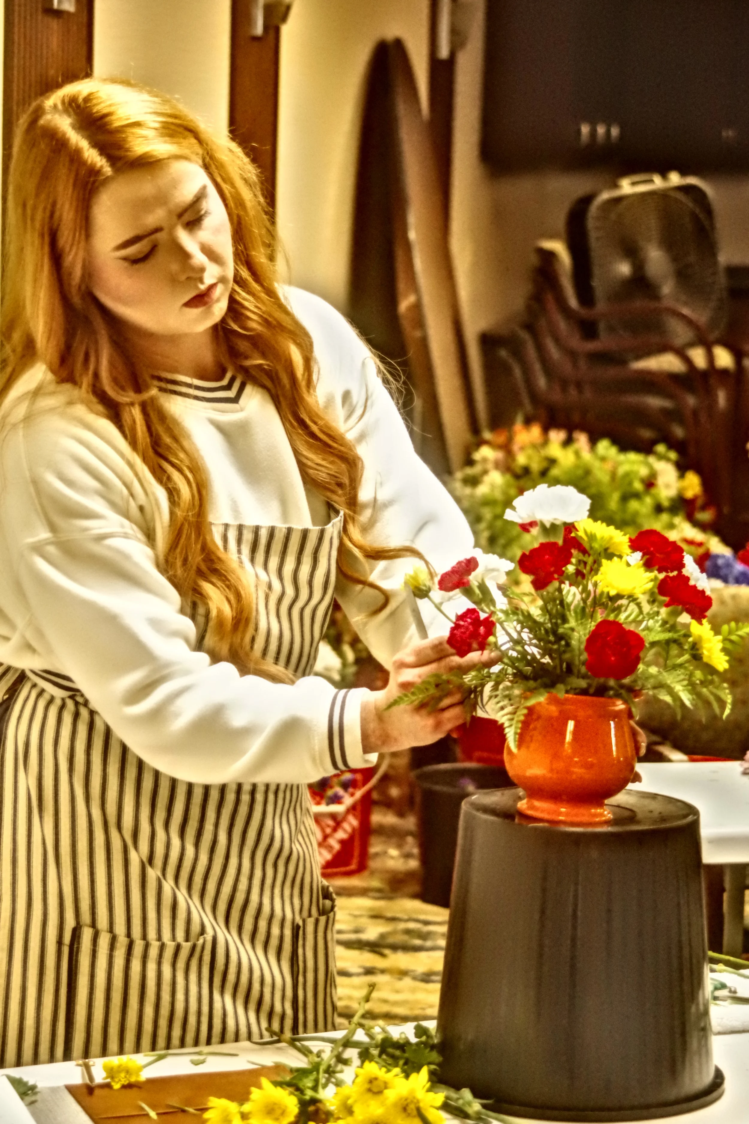 A woman with long red hair arranging flowers in a vase. She is wearing a striped apron over a sweatshirt and standing in a room with chairs and a fan in the background.
