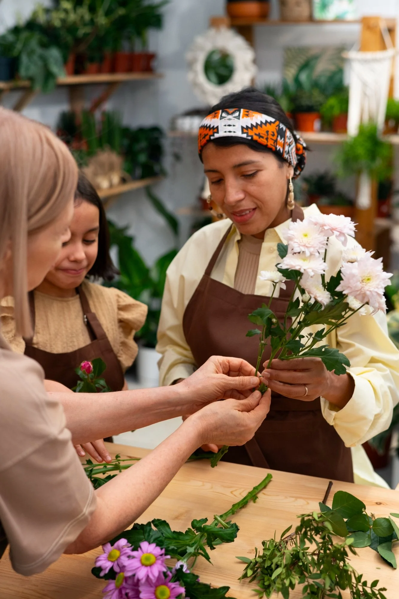 Four women gather around a table in a flower arranging workshop, holding and examining pink and white flowers and green foliage among various flowers and greenery on the table.