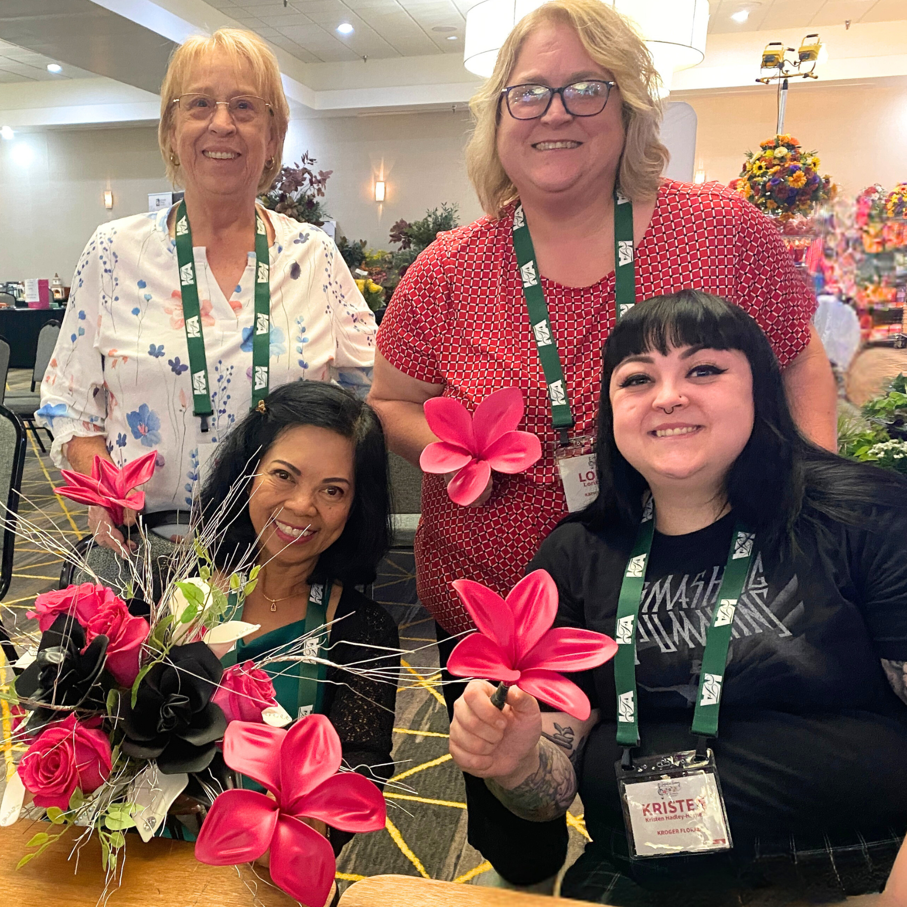 A group of women posing at a floral event, with flowers and plants visible in the background. One woman in front is holding a pink flower, and a woman to her right is smiling with a red and white patterned top. They are all wearing conference name tags.