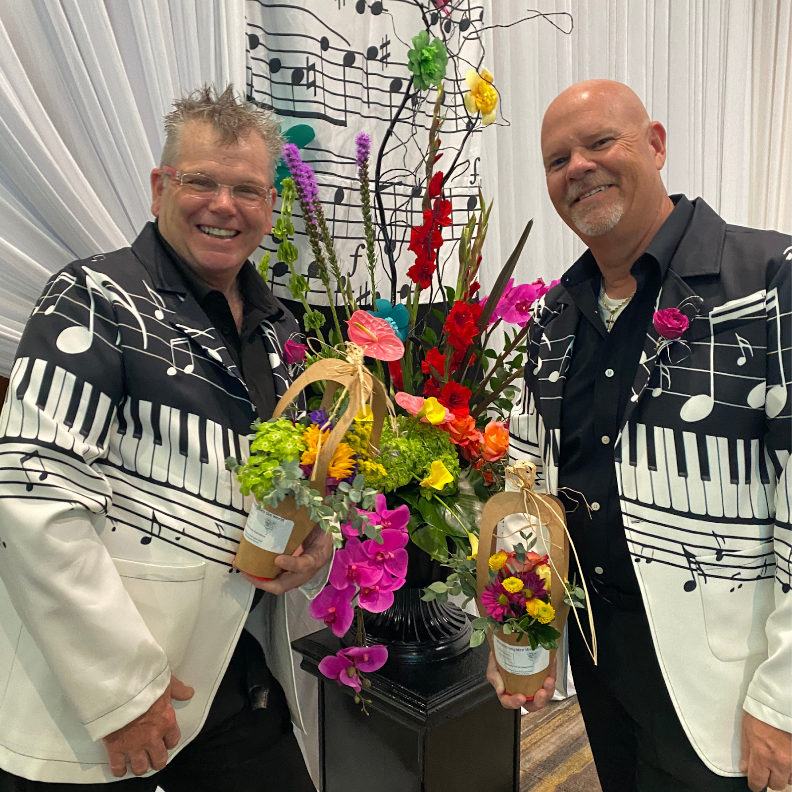 Two men dressed in black and white musical note themed suits standing in front of a flower arrangement, smiling at the camera. One man is holding a small potted flower, and the background features a wall with decorative muscial notes and lines.