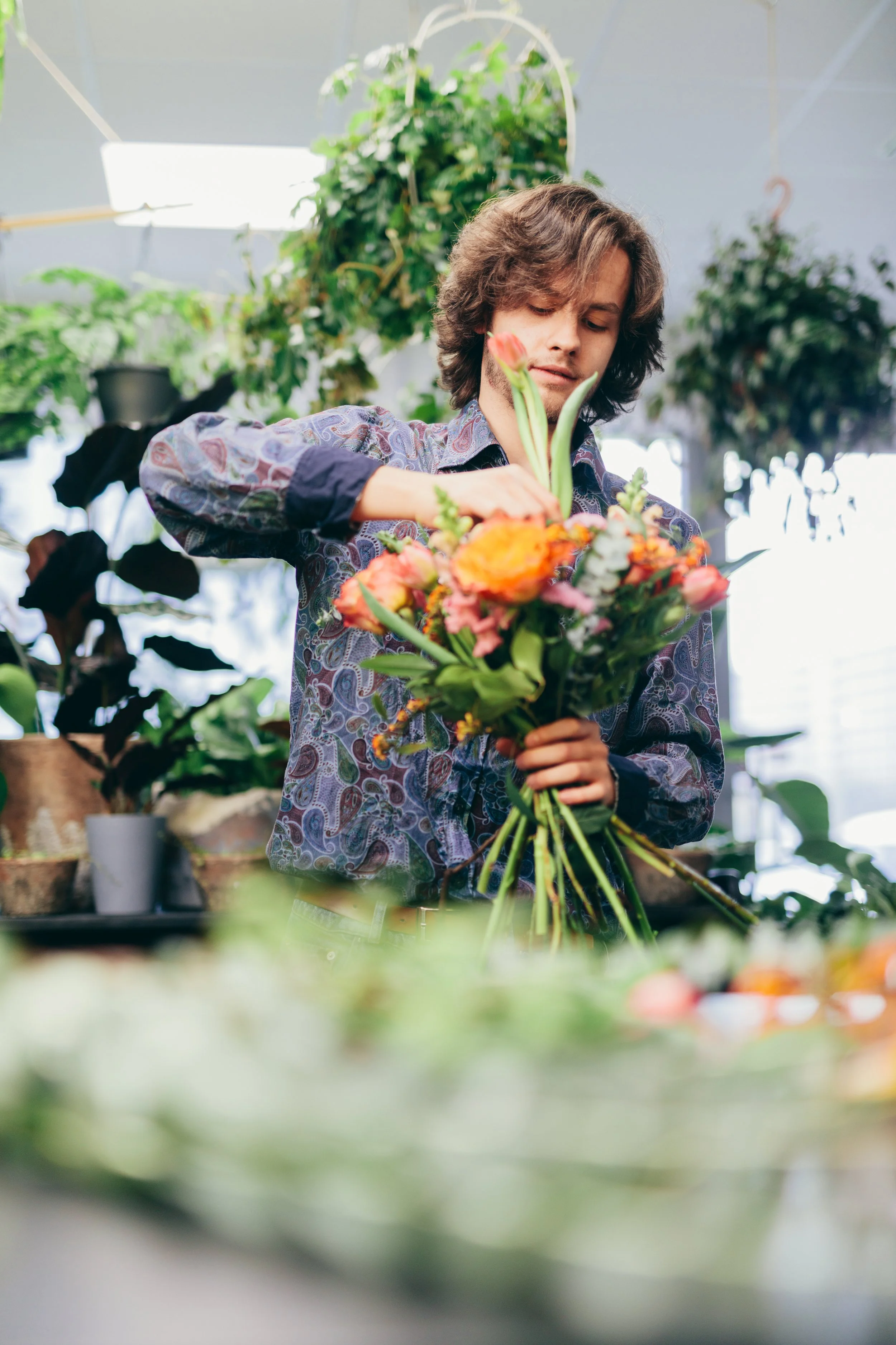 A young man with wavy brown hair and light beard arranging a bouquet of flowers in a greenhouse surrounded by green plants.