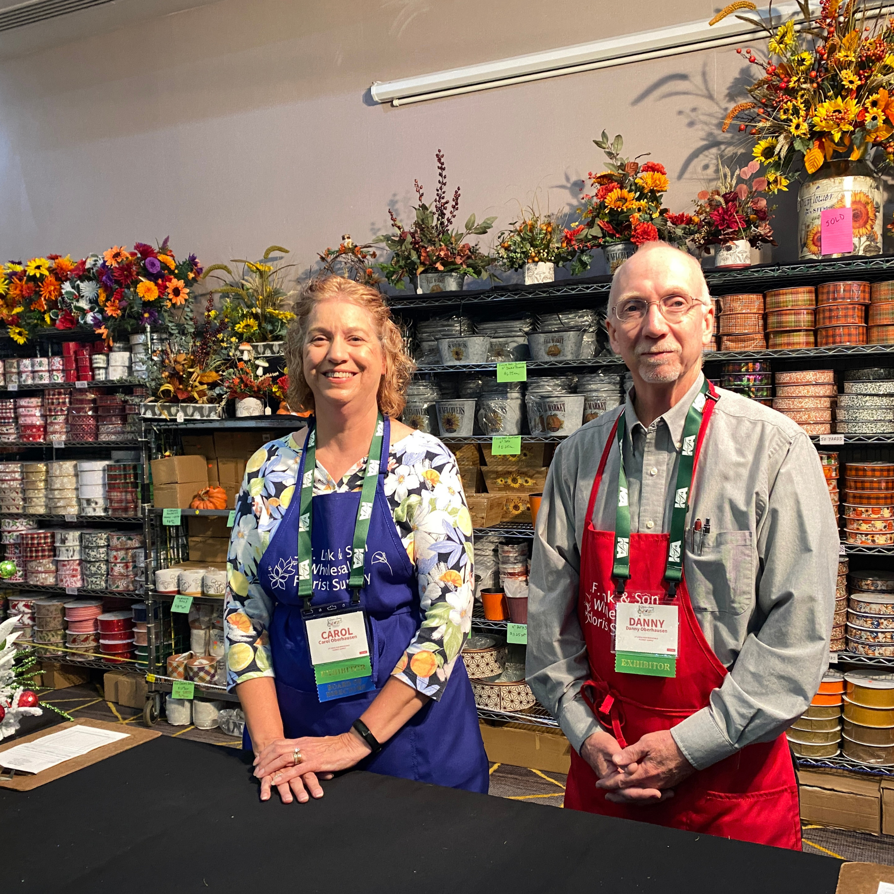 A woman and a man standing in a store with shelves of decorative pots, flower arrangements, and gift wrap behind them. The woman is smiling and wearing a floral print shirt and a blue apron, and the man is wearing glasses, a gray shirt, and a red apron. Both have name tags and are smiling at the camera.