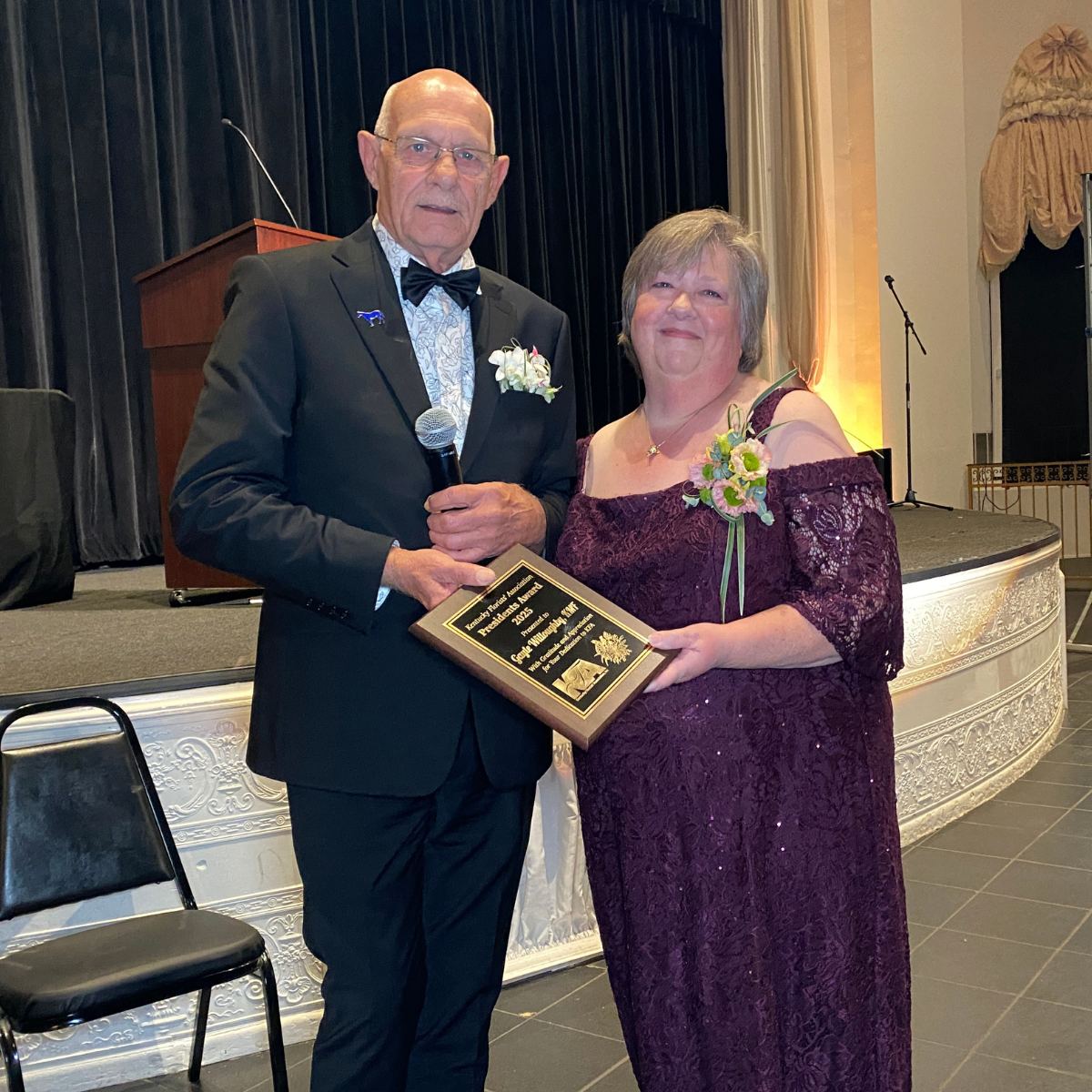A man and woman standing together on stage at an awards ceremony, holding a plaque and smiling. The man wears a black tuxedo with a bow tie and glasses, and the woman wears a burgundy lace dress with floral corsages.