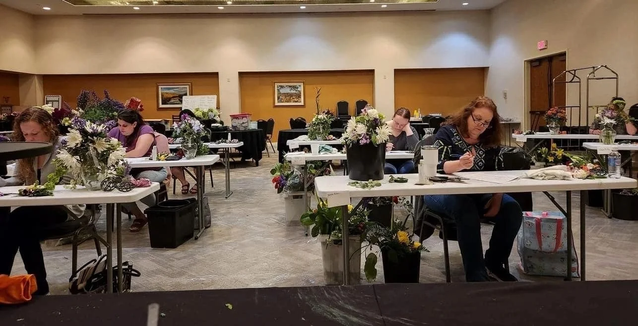 Women sitting at tables with flower arrangements, working on flower arrangements in a spacious indoor room.