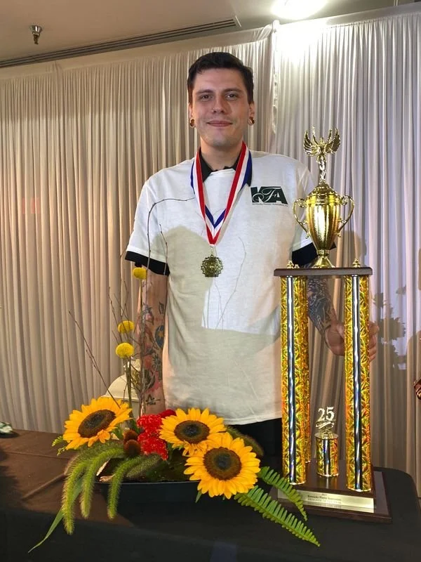 A young person standing behind a table with a large trophy, wearing a white sports jersey with a logo, a gold medal around their neck, and sporting tattooed arms. There are yellow sunflowers and other flowers in front of the table.