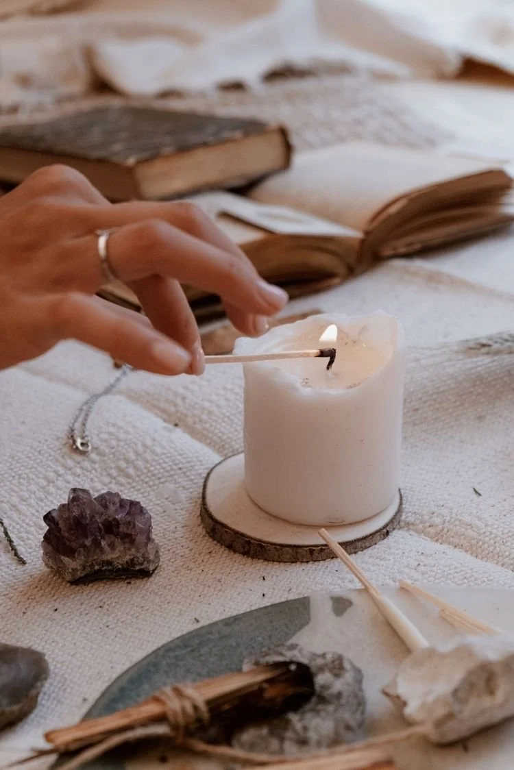 Person lighting a white candle with a match on a table decorated with crystals and open books.