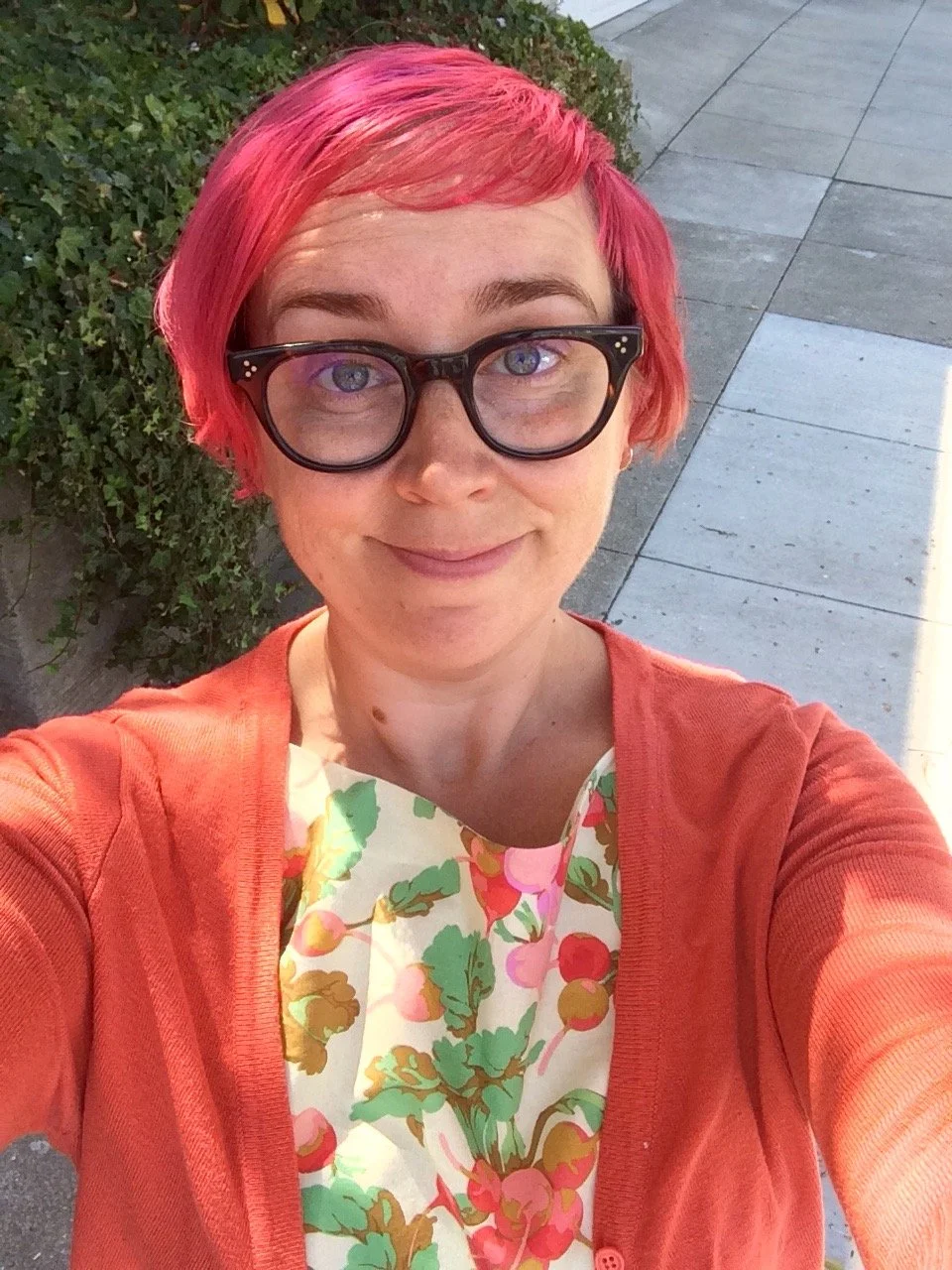 An avid sewist with short pink hair and glasses taking a selfie outdoors, wearing a floral dress she sewed herself and an orange cardigan.