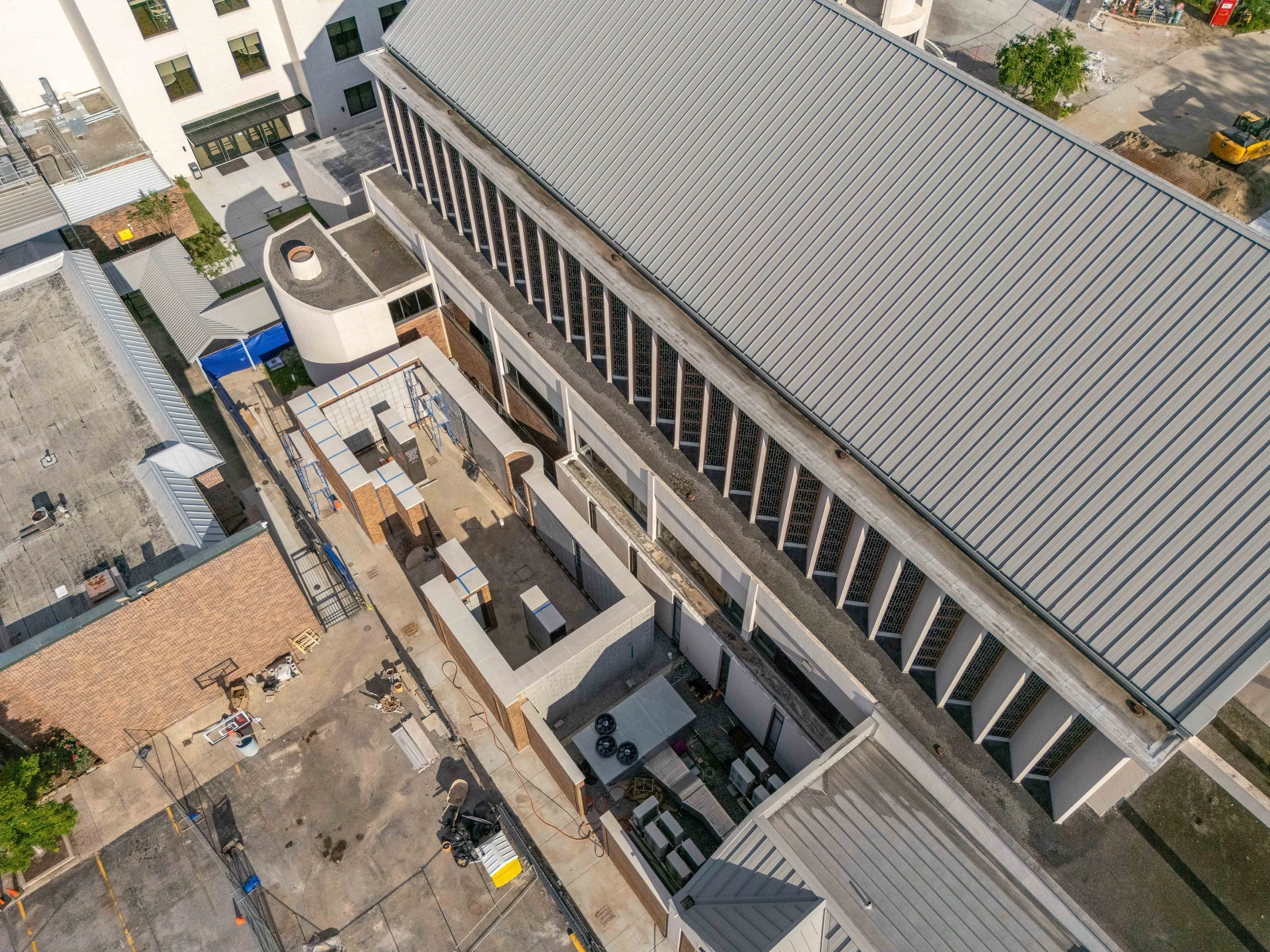 Aerial view of a large commercial building with a metal roof, adjacent to a smaller brick building with various equipment, and outdoor areas with construction materials and machinery.
