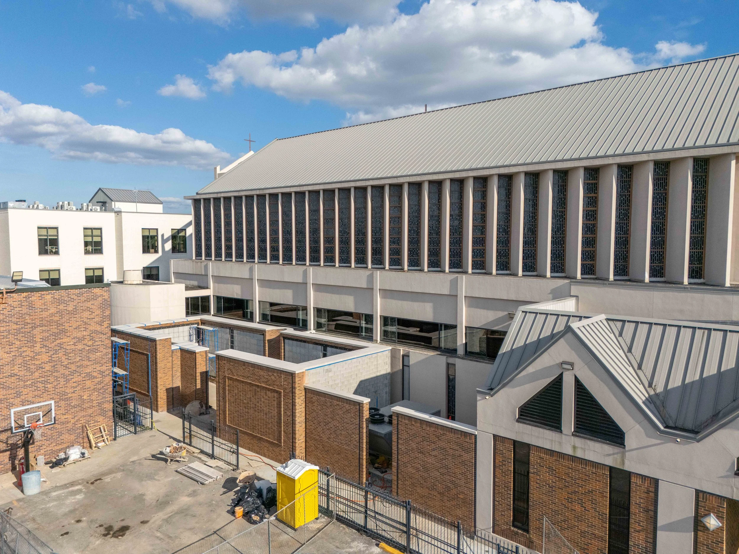 Construction site of a modern church building with a large gray metal roof, brick walls, and tall windows. There is ongoing work with construction materials and equipment visible, surrounded by a metal fence. The sky is partly cloudy with blue sky.