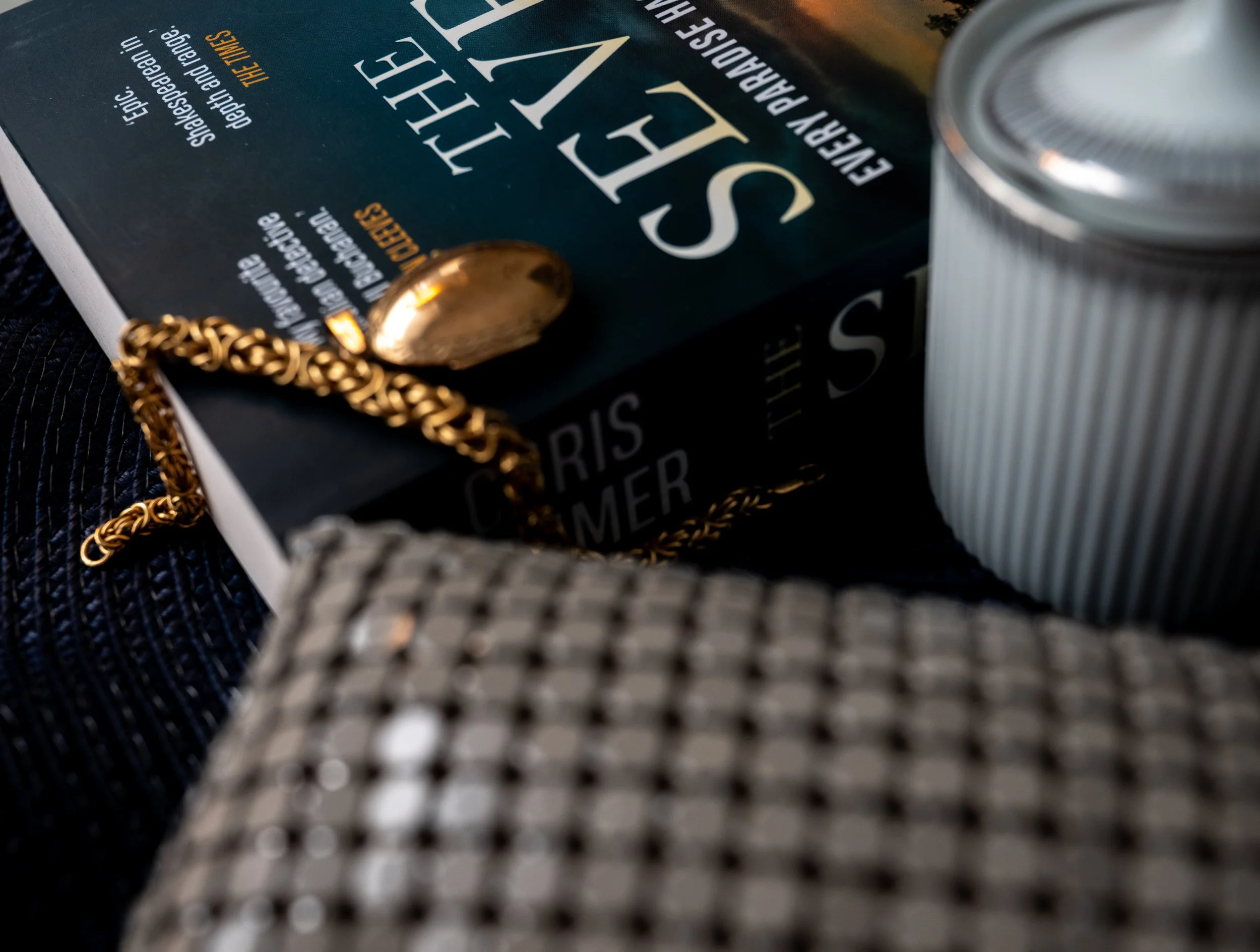 Close-up of a book titled 'The Seven' by Chris Hammer, a gold chain necklace with a pendant, a small sugar container with a metallic lid, and a textured fabric surface.