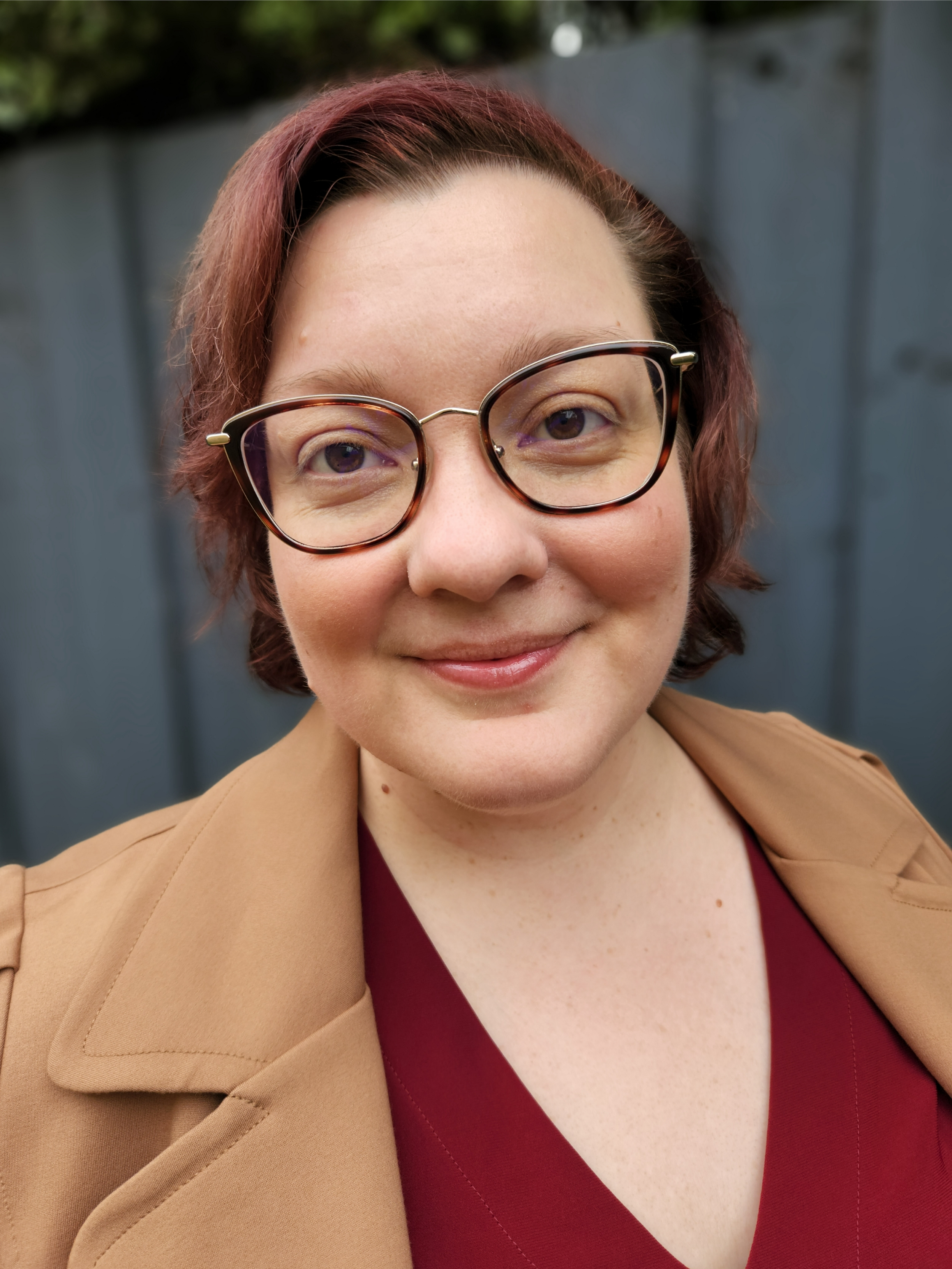 A photo of Kristen Duthie with short reddish-brown hair, glasses, and a friendly smile, wearing a tan coat over a burgundy top, standing outdoors in front of a grey fence.
