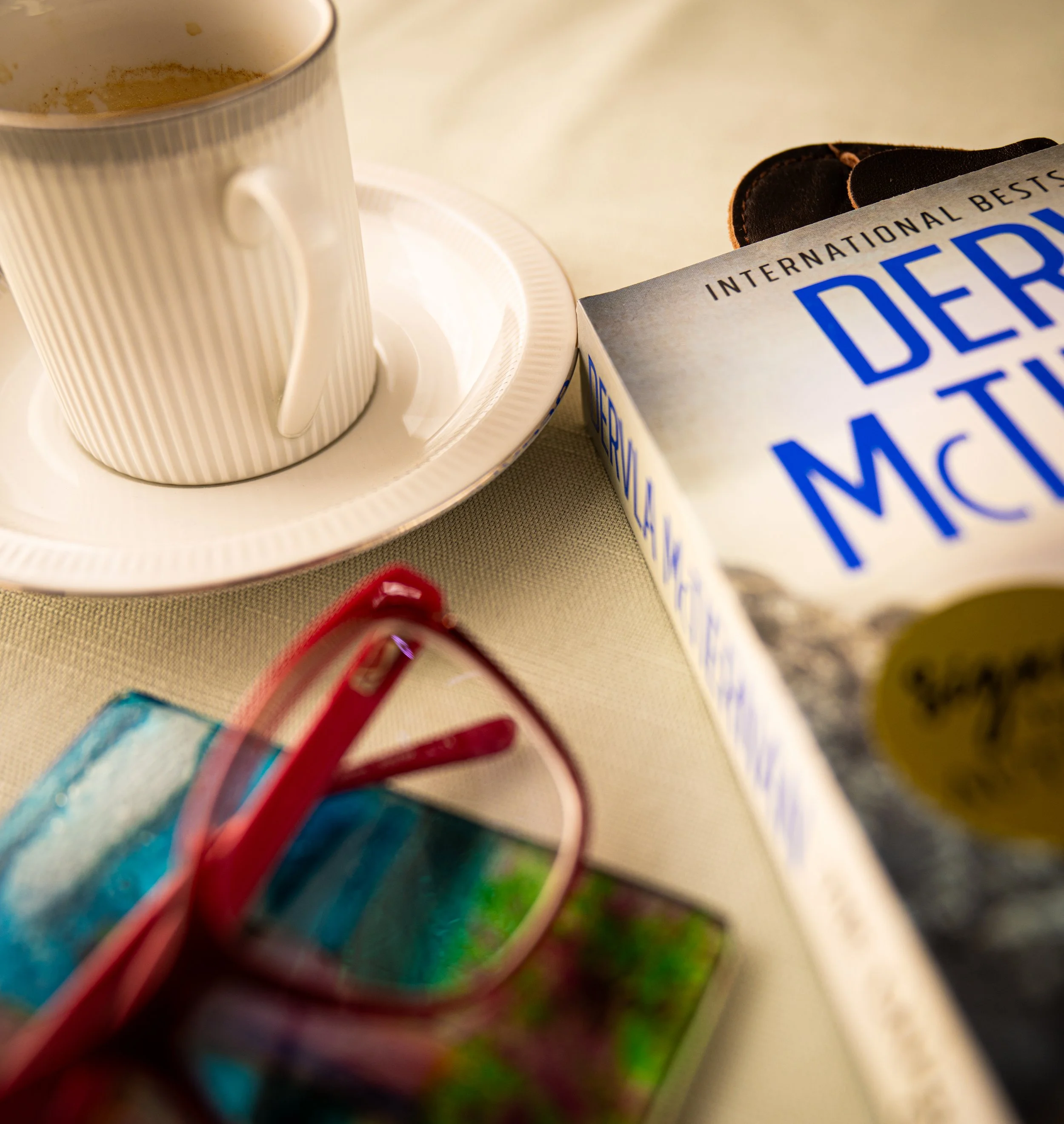 A white coffee mug on a saucer next to a box of deep-minted chocolates and a pair of pink sunglasses with reflective lenses on a beige surface.