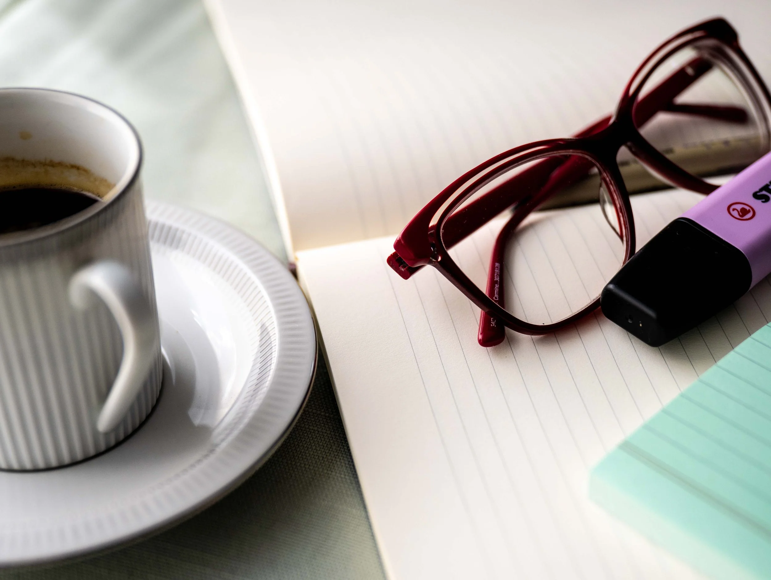 Red eyeglasses, a purple highlighter, an open notebook, a cup of black coffee on a saucer, and a stack of colored paper on a table.