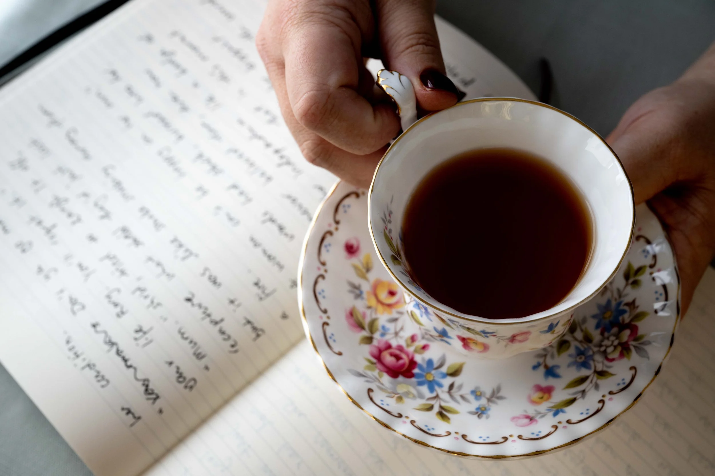 A person holding a floral teacup filled with tea above a handwritten journal or notebook.