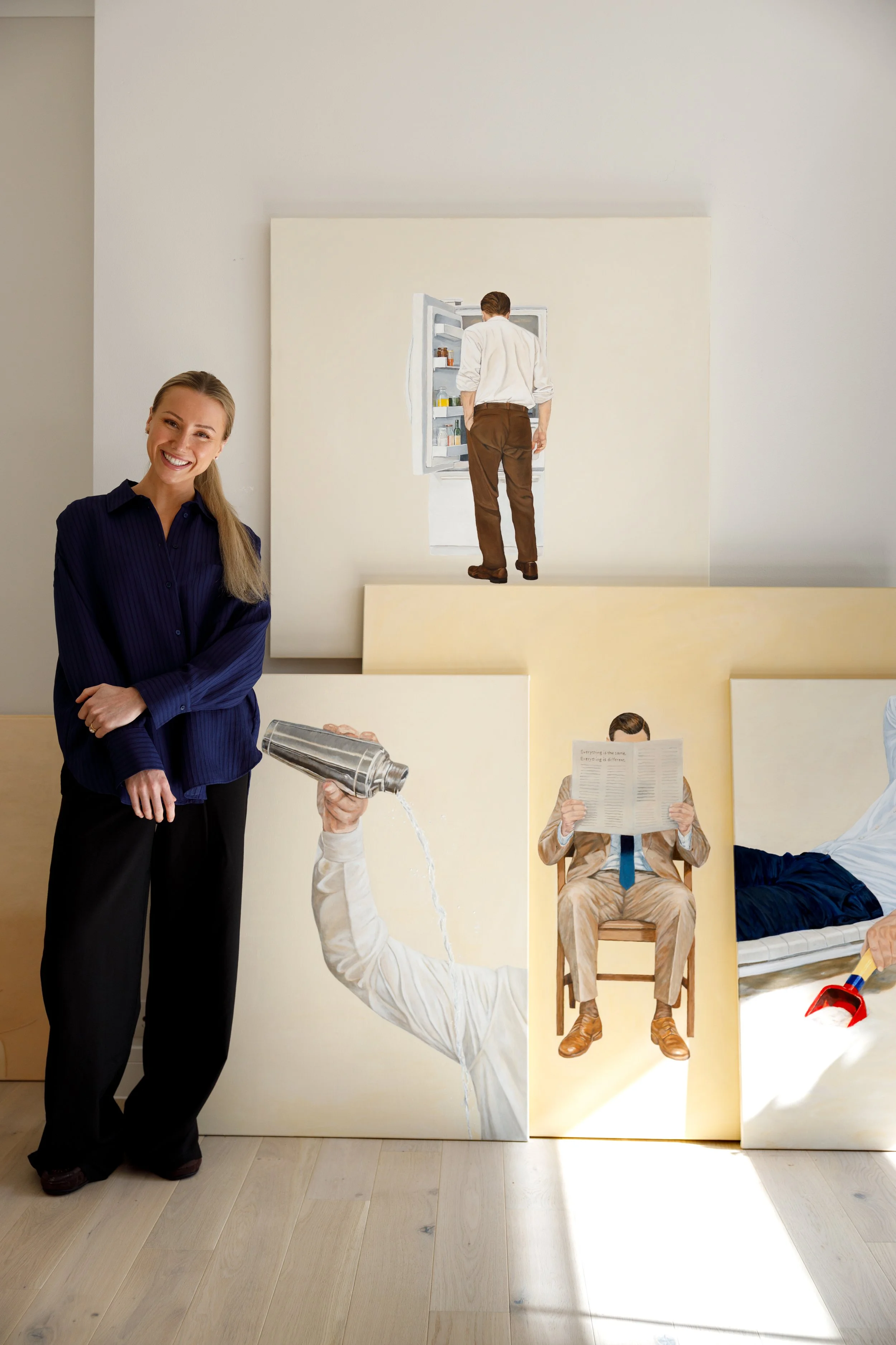 A woman standing next to four paintings, three of which depict people involved in various activities, and one of which is a sleeping person on a bed. The woman is smiling and holding a bottle of water.