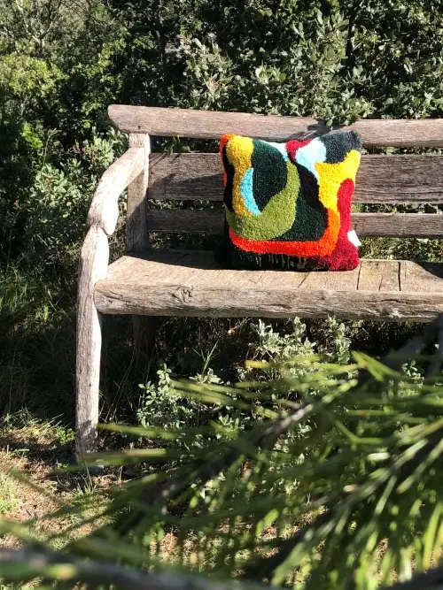 Un banc en bois avec coussin coloré de la Maison et Atelier Ama Adao créé par Chloé Fortunet situé en plein air en Provence avec des arbres et des plantes en arrière-plan.