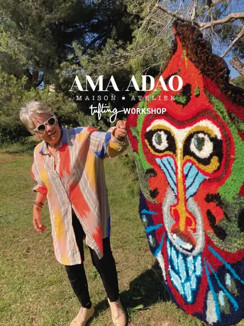 Une femme âgée avec des lunettes blanches, portant une chemise colorée, pose à côté d'une sculpture en tufting de singe faite par la Maison et Atelier Ama Adao créé par Chloé Fortunet, en plein air en Provence avec des arbres en arrière-plan.