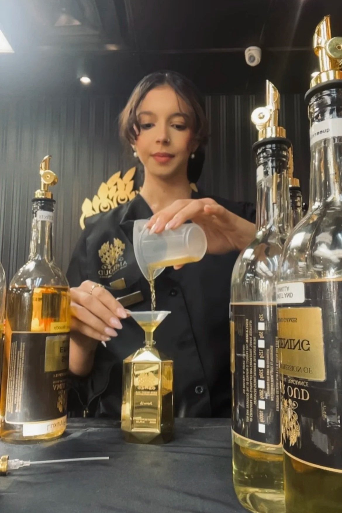 A woman in black attire pouring a golden-colored liquid into a small glass, with several large bottles of the same liquid on the table in front of her, indoors with black walls and gold decorative elements.