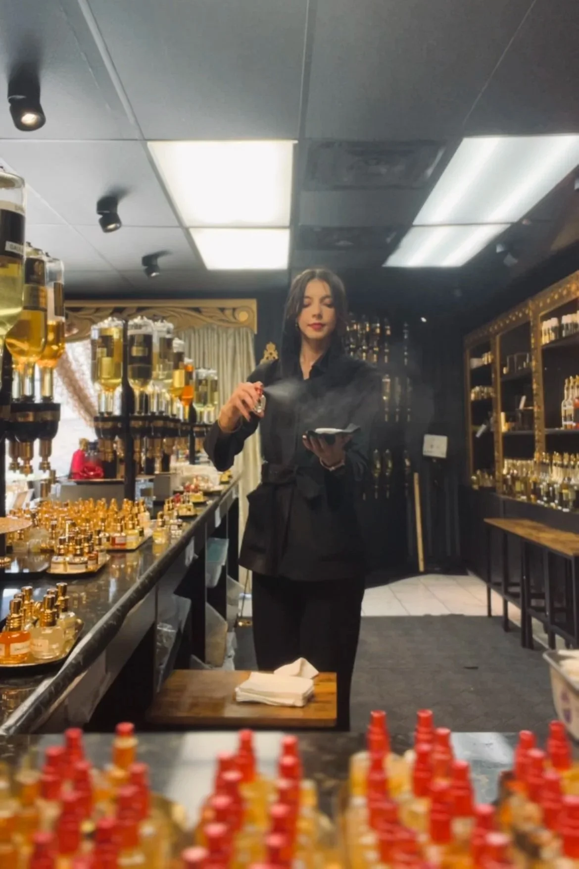 A woman with dark hair and red lipstick standing in a perfume shop, holding a perfume bottle and a mobile phone, surrounded by shelves filled with perfume bottles.