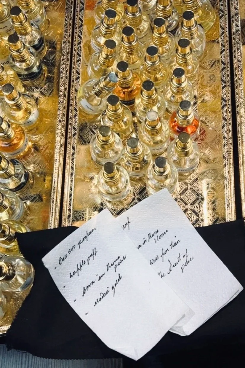 A display of multiple bottles of perfume or cologne, arranged on a decorative gold and black patterned surface. In front, there are handwritten notes on white paper, resting on a folded black cloth.