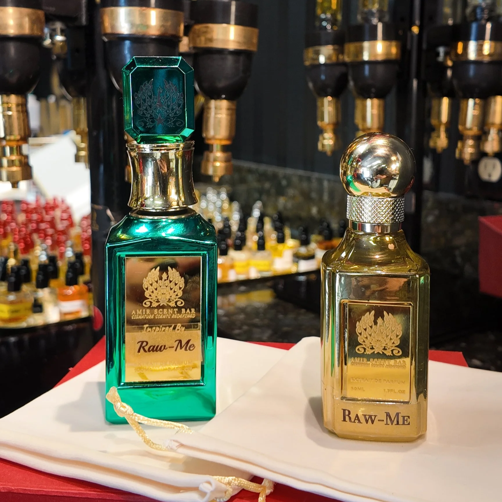 Two bottles of perfume on a white cloth, with a background of shelves filled with small bottles. The left bottle is teal with a gold label and cap, labeled 'Raw-Me,' and the right bottle is gold with a silver cap, also labeled 'Raw-Me.'