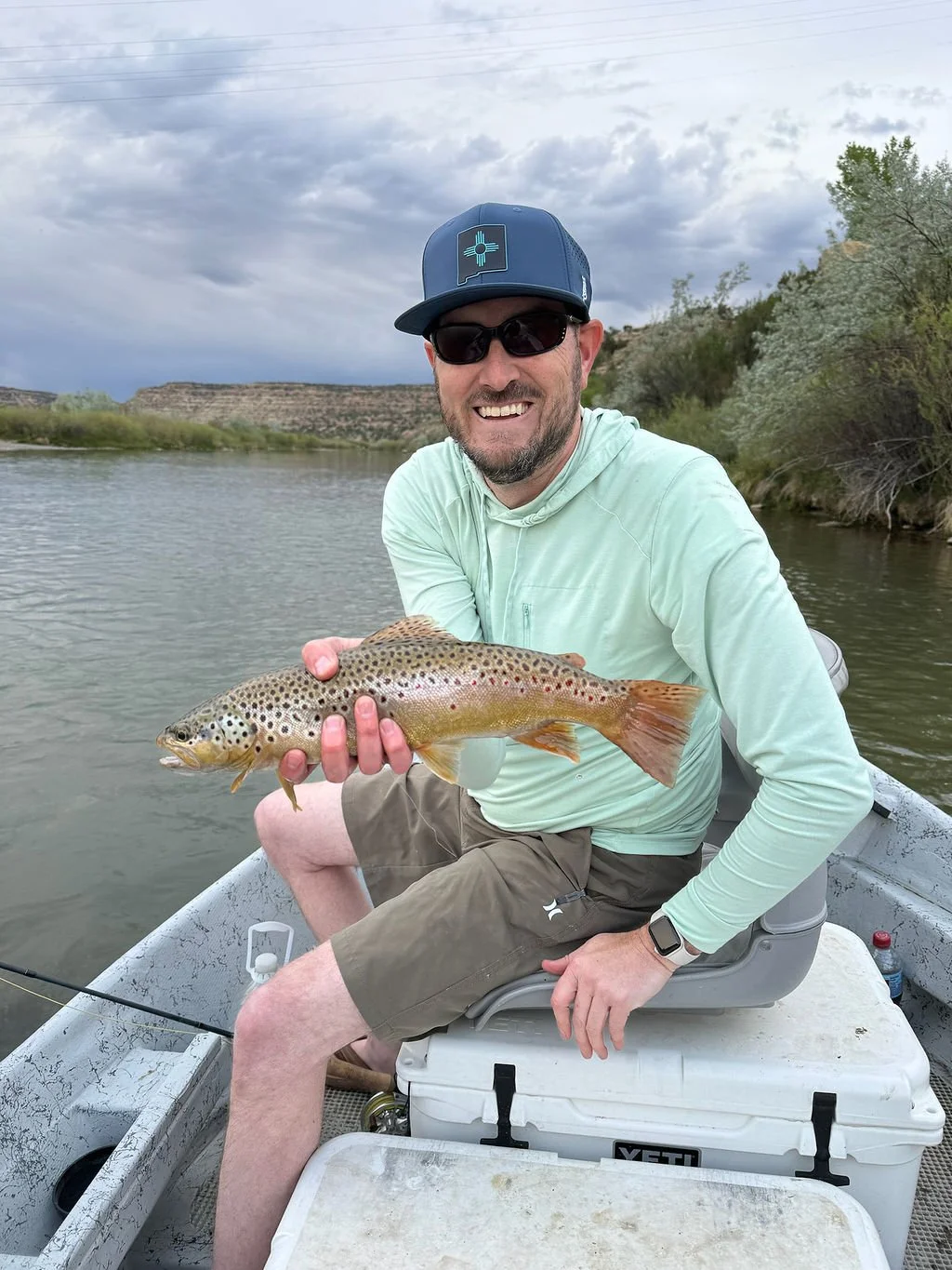 A man wearing sunglasses and a cap sitting in a boat on a river, holding a large fish with a colorful spotted body, smiling at the camera.