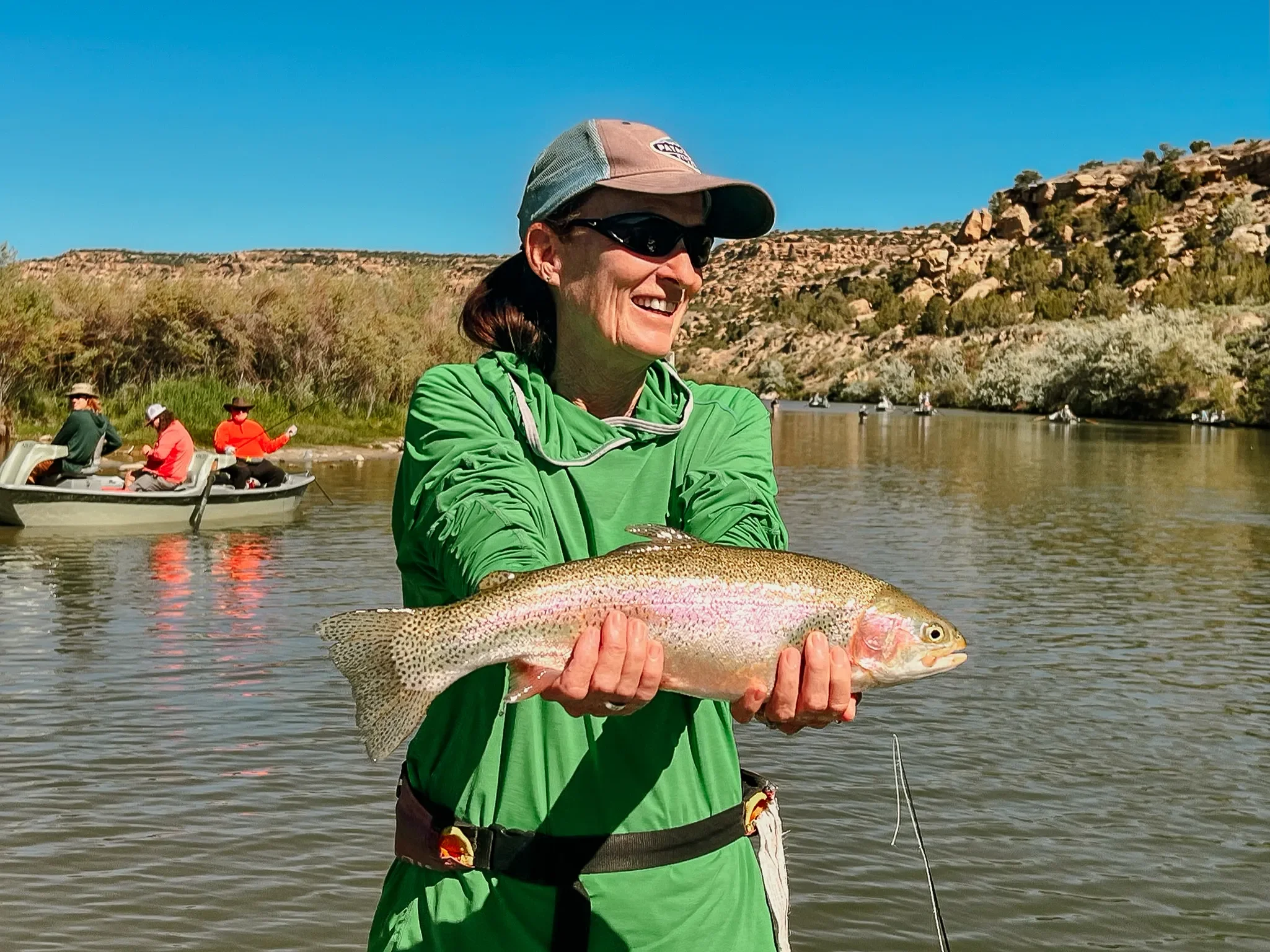 A woman in a green jacket holding a large fish she caught in a river, with a background of rocky hills and other people in boats.
