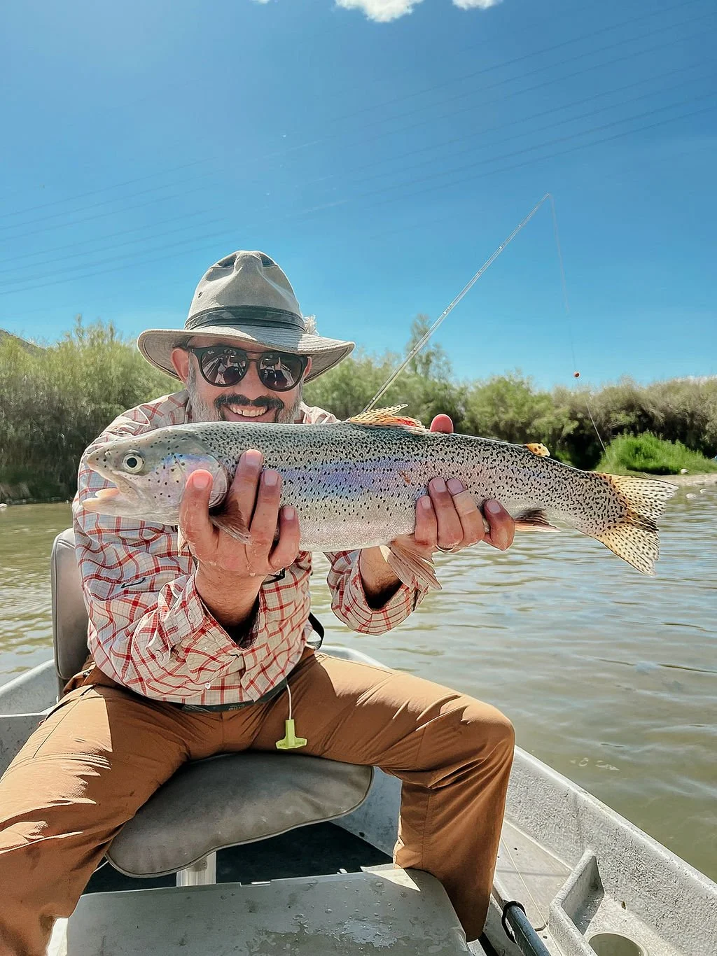 A man wearing a wide-brimmed hat, sunglasses, and a checkered shirt smiling while holding a large rainbow trout on a boat in a river.