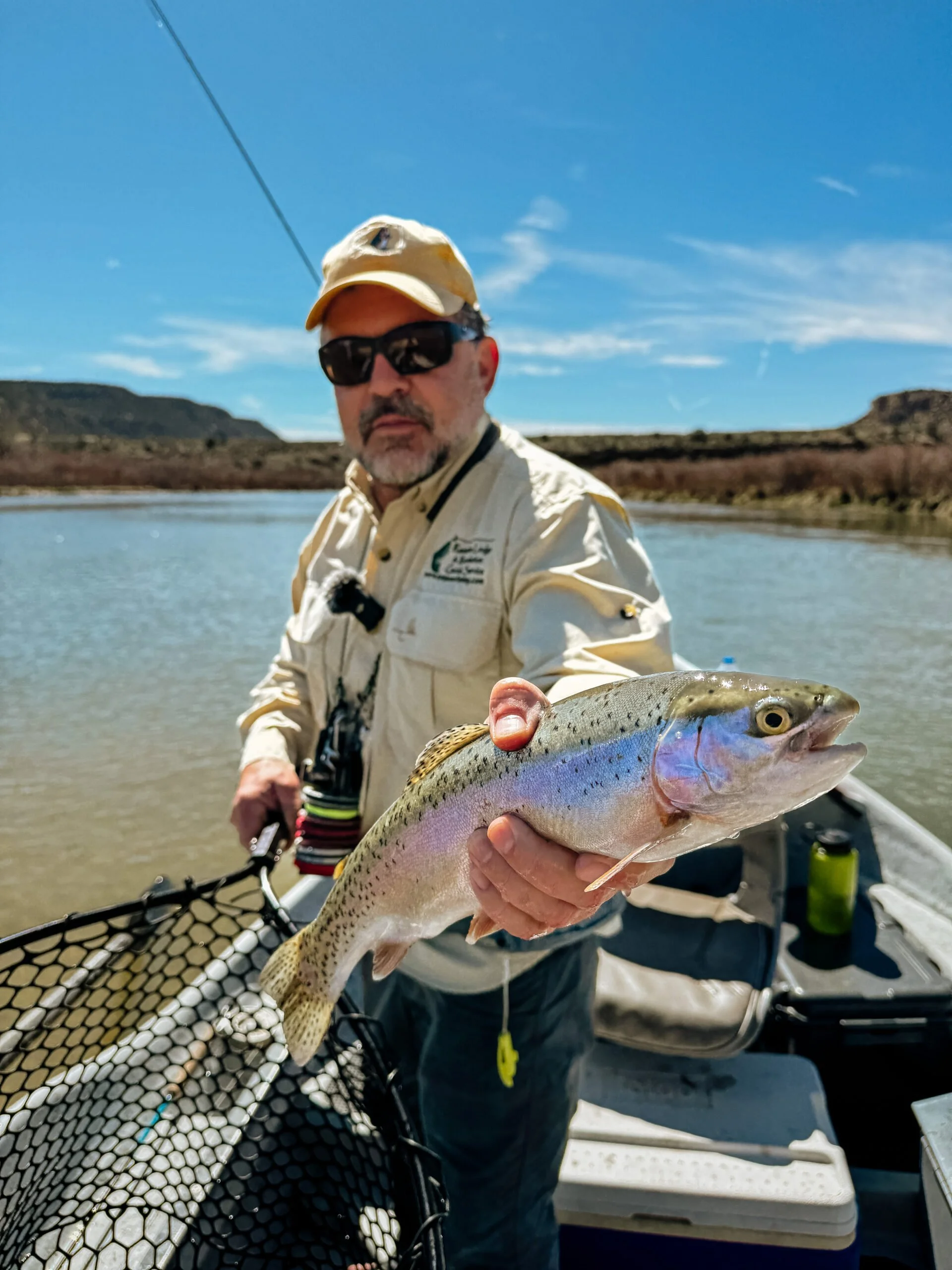 A man wearing sunglasses, a beige hat, and a light-colored shirt standing on a boat, holding a rainbow trout he caught, with water and a mountainous landscape in the background.