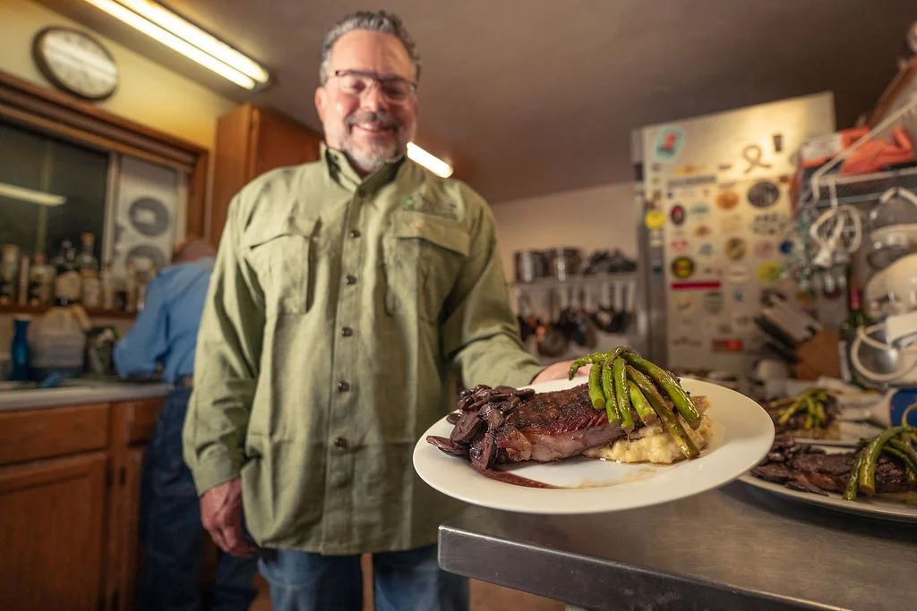 Smiling man holding a plate with a cooked steak topped with green beans in a rustic kitchen.