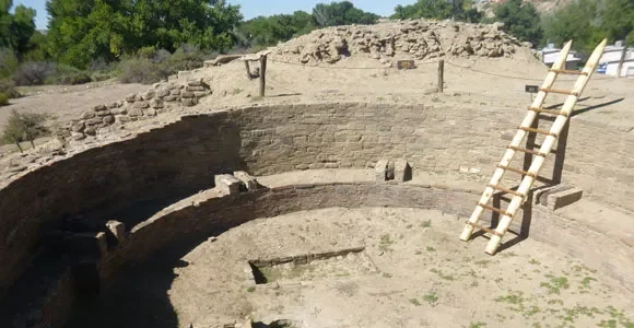 An archaeological excavation site with circular stone walls and a wooden ladder leaning against the outer wall.