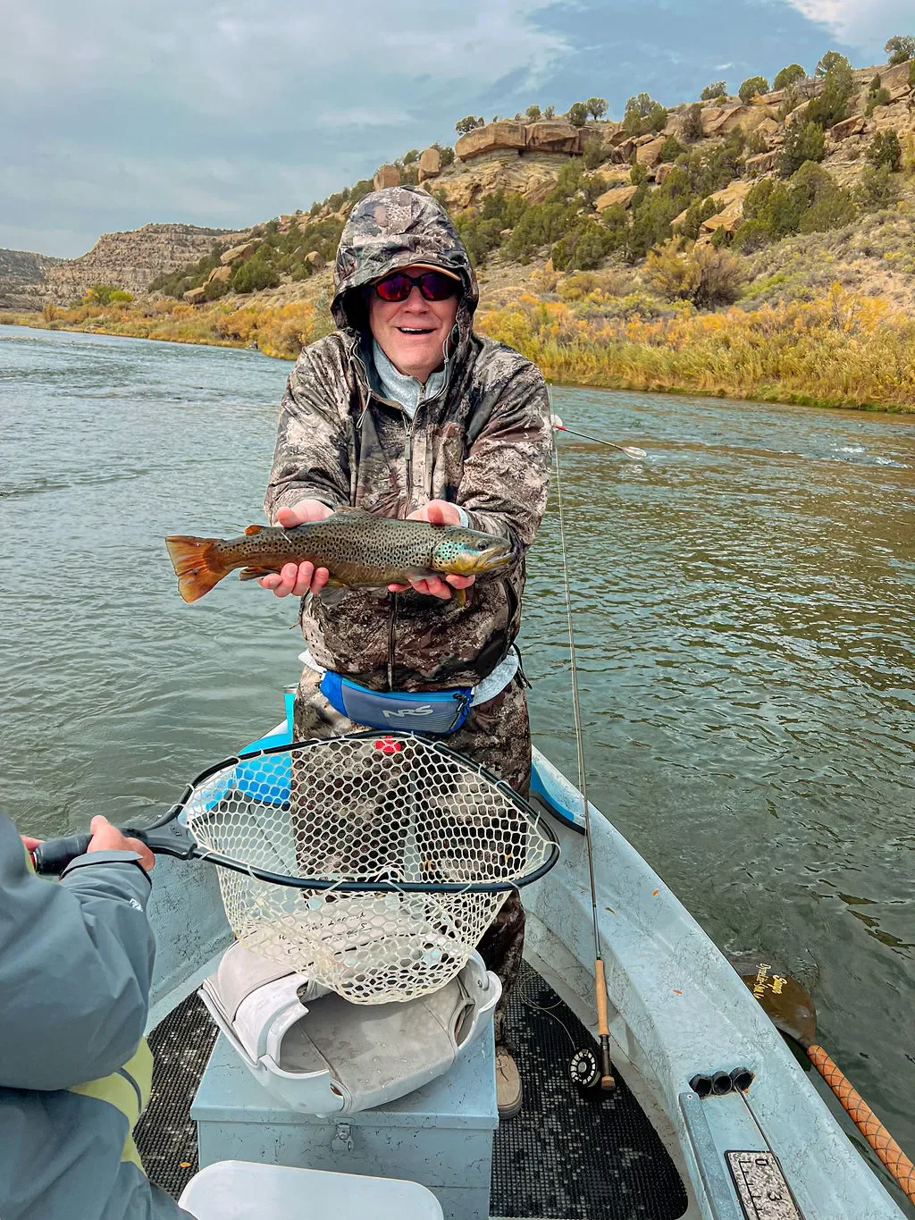 Person in camouflage outfit and sunglasses holds a rainbow trout on a boat on a river with rocky hills and sparse trees in the background.