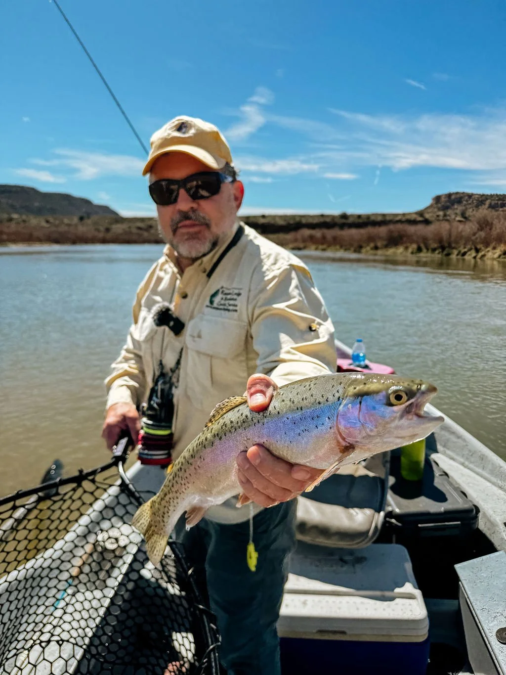 Man in sunglasses and a beige hat holding a fish on a boat on a river with hills in the background.