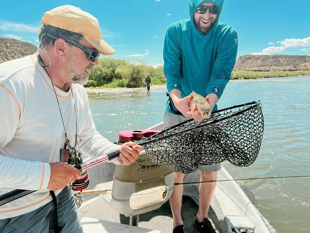 Two men on a boat, one older with a gray beard and sunglasses holding a fishing net, and a younger man with a hood, sunglasses, and a beard holding a fish. They are on a river with hills and trees in the background.