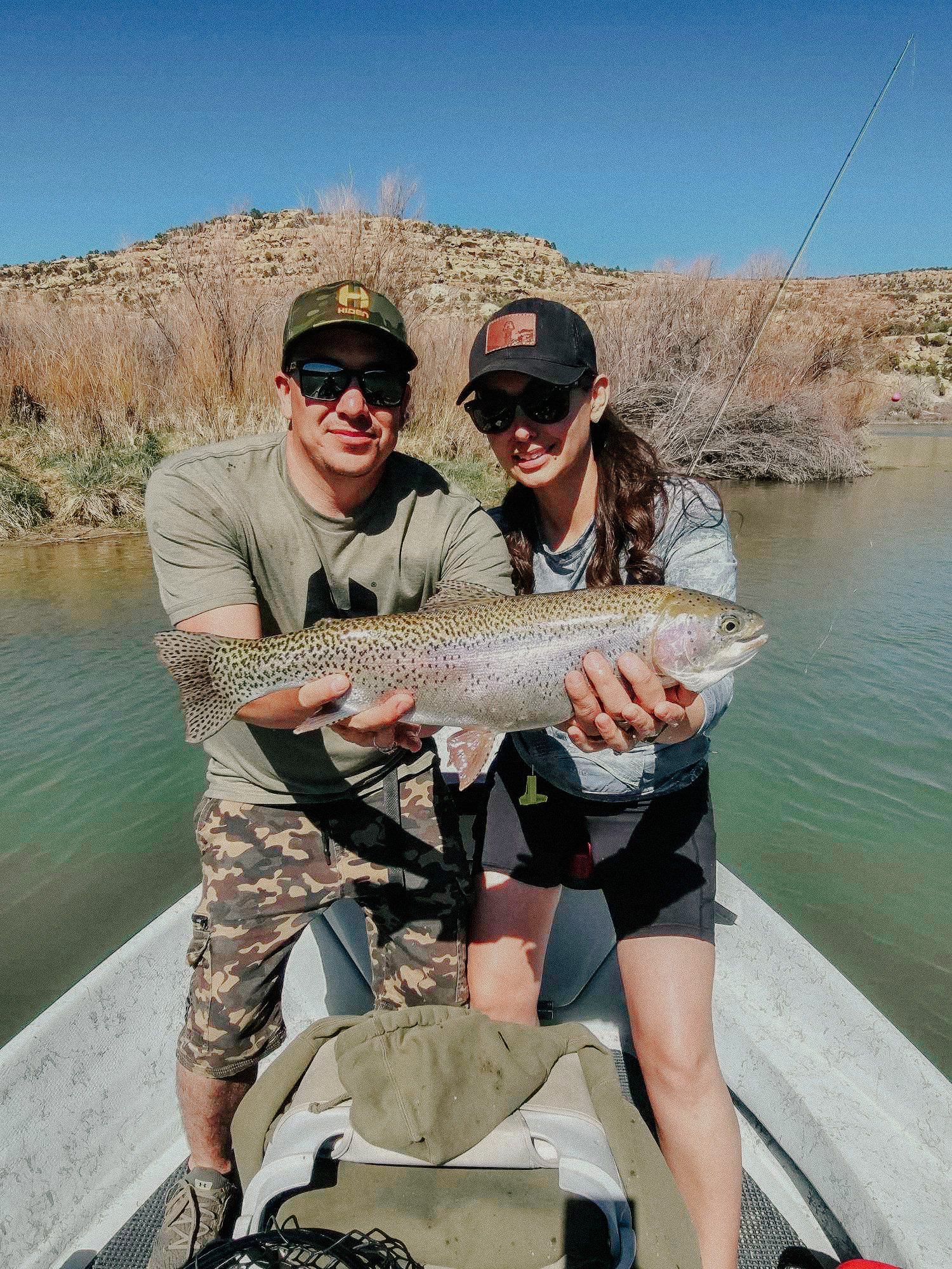 Two people on a boat holding a large fish with a mountainous landscape background.