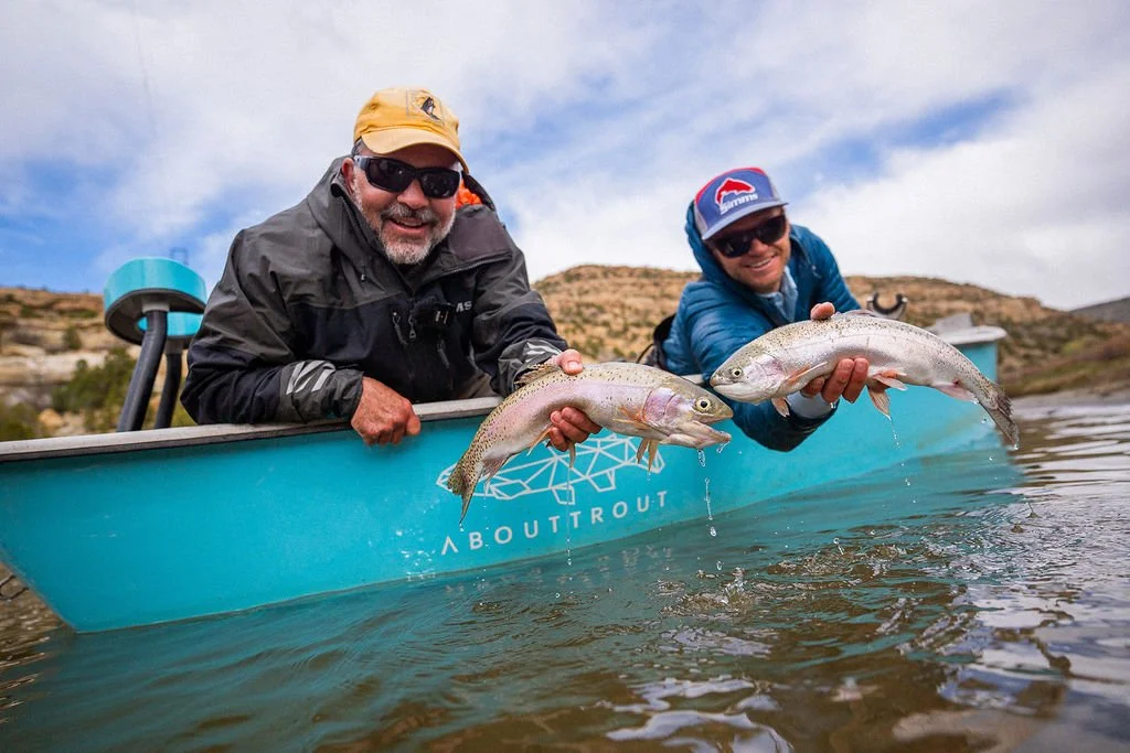 Two men in a blue boat holding fish they caught, with a desert landscape in the background.