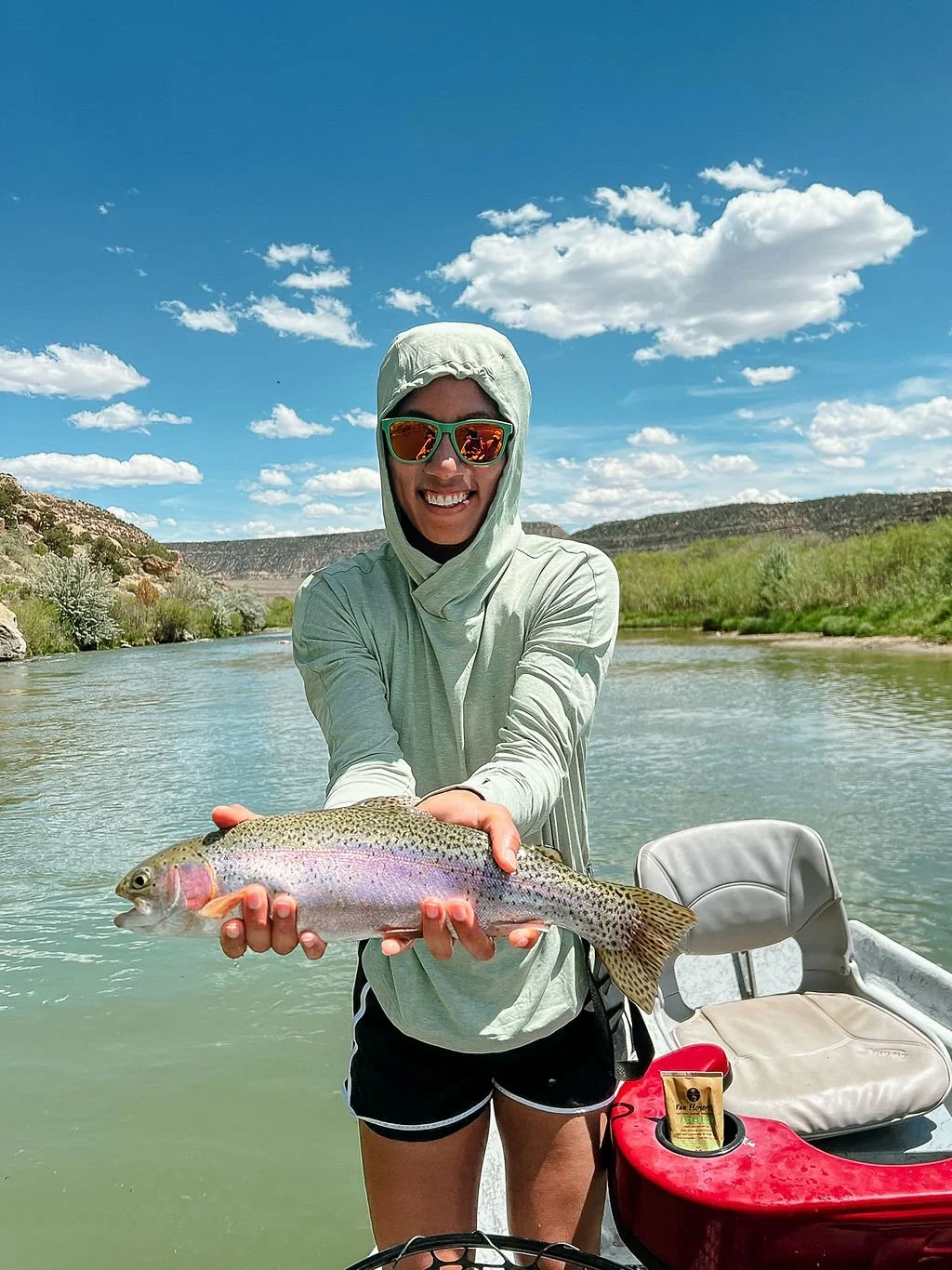 A woman in sunglasses and a hooded long sleeve shirt holds a rainbow trout on a boat in a river with green hills and a blue sky with clouds in the background.
