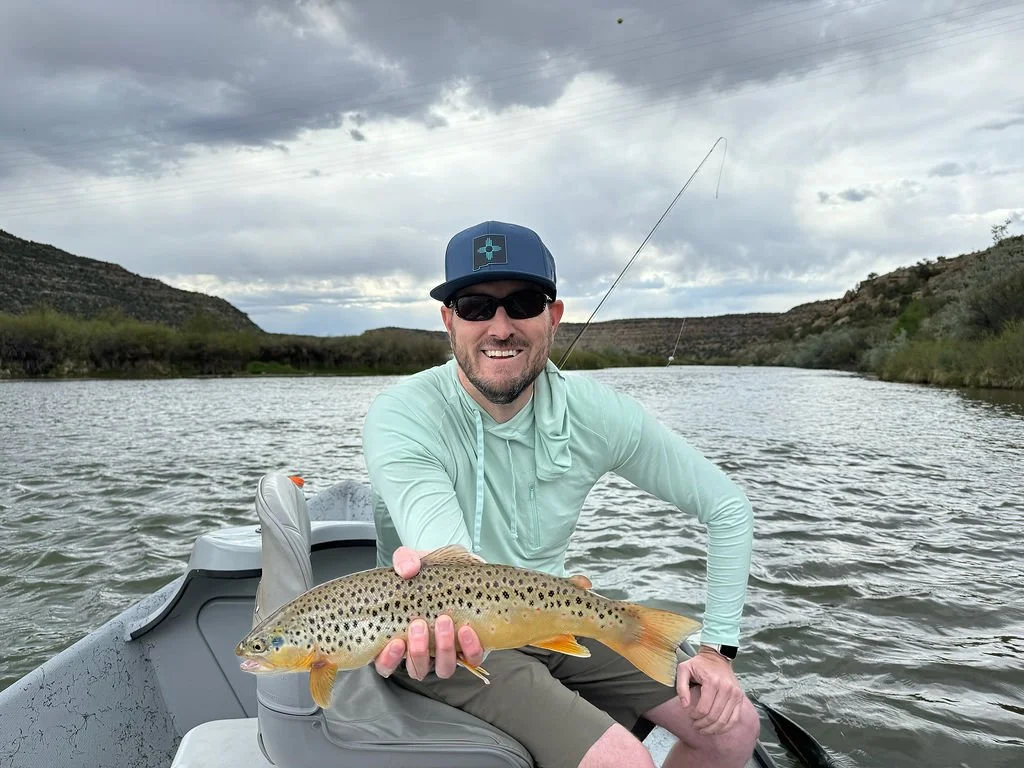 A man in sunglasses and a blue cap, sitting in a boat on a river, holding a caught fish with a view of riverbanks and cloudy sky in the background.