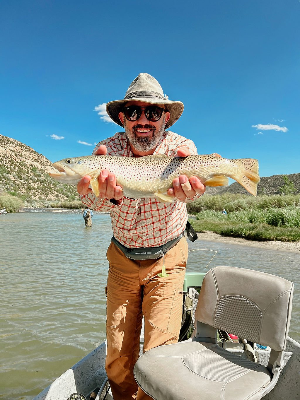 Man in a hat and sunglasses holding a large fish on a boat by a river, with hills and blue sky in the background.