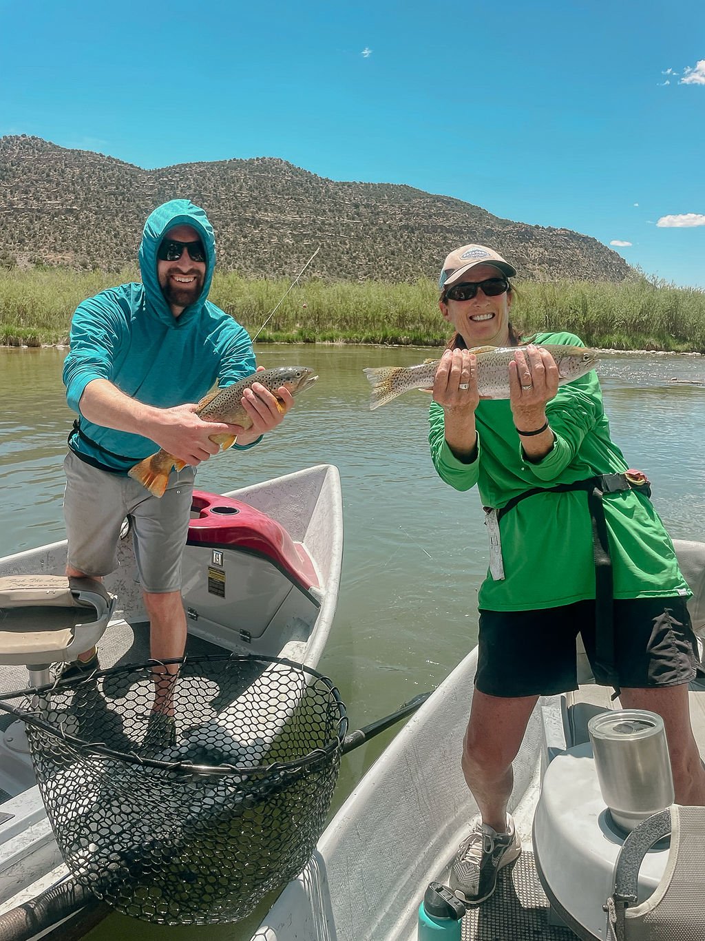Two people on a boat holding fish they caught, with a mountain and blue sky in the background.