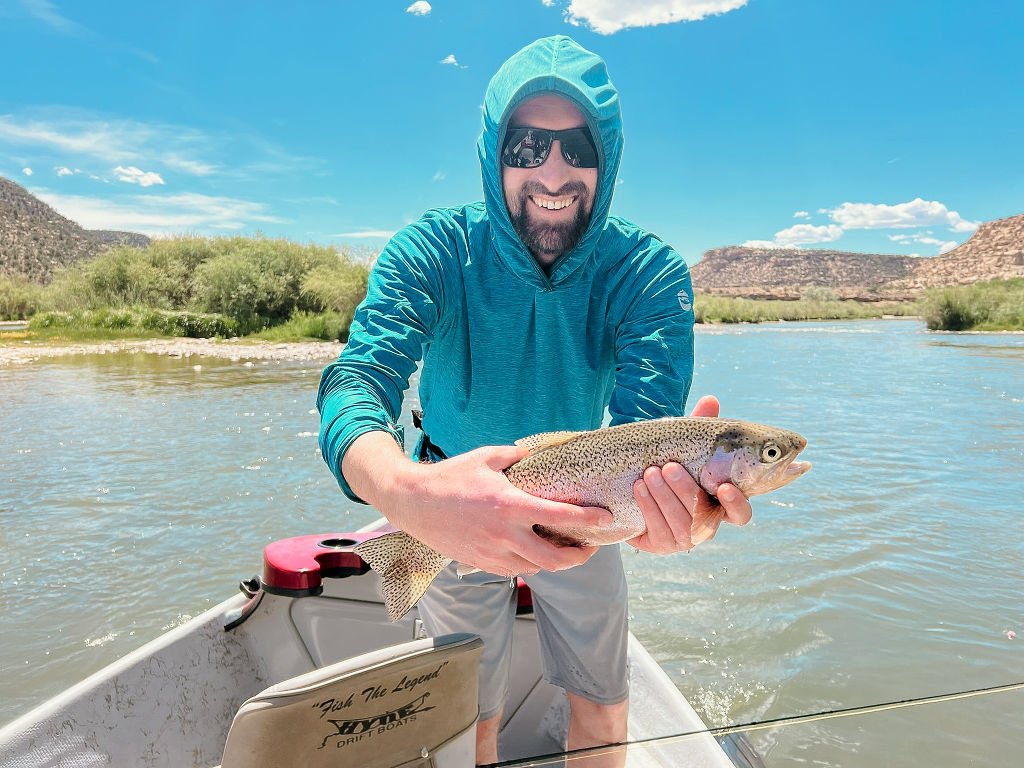 A man in outdoor fishing gear holding a rainbow trout on a boat with a river and desert landscape in the background.