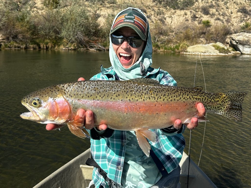 Person in outdoor clothing and sunglasses holding a large rainbow trout fish on a boat in a river, with a rocky and shrubbery background.