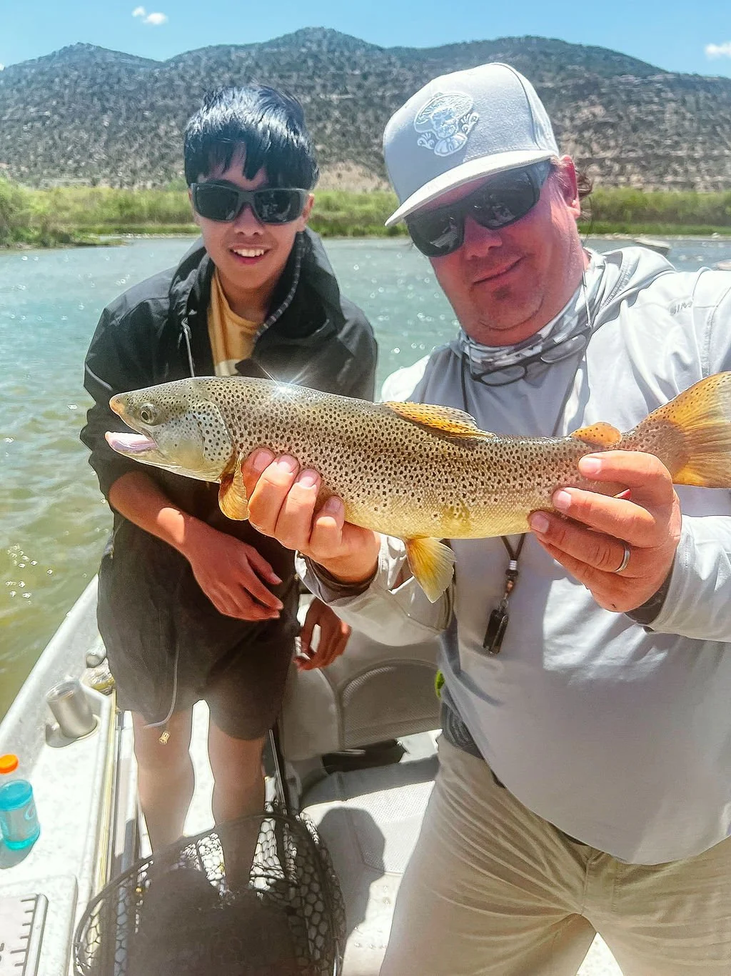 Two men posing on a boat with a large rainbow trout, outdoors near a river with mountains in the background.