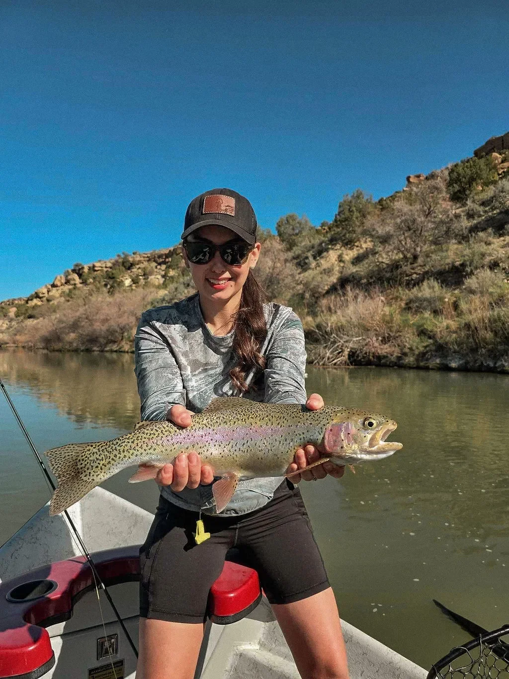 Woman in sunglasses and hat holding a large rainbow trout in a boat on a river with hillside and clear blue sky in the background.