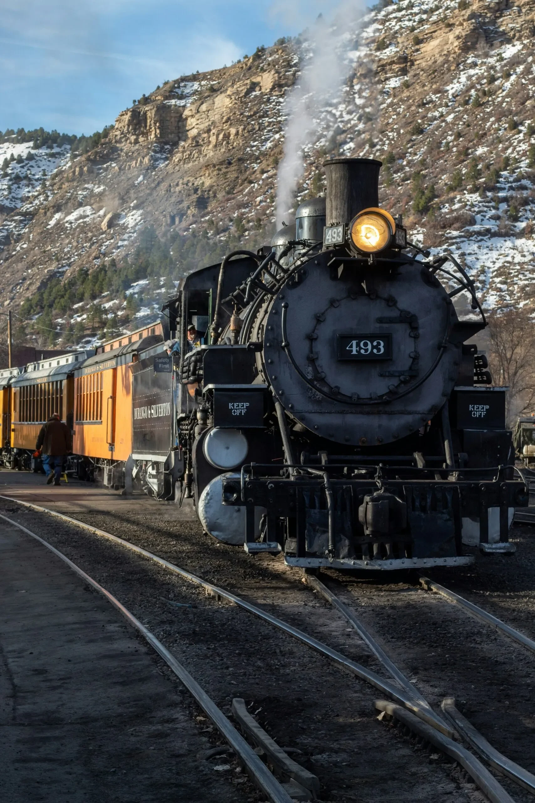 A vintage steam train moving along railway tracks with mountains covered in snow in the background.